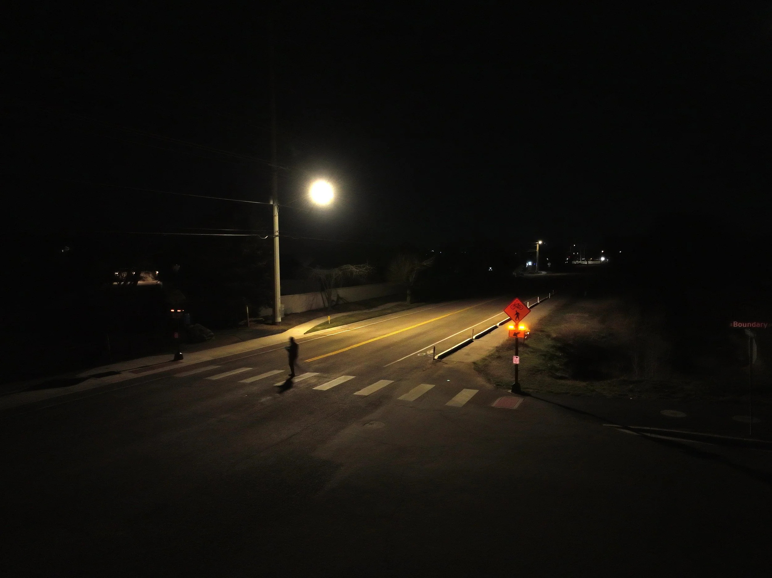 Nighttime street scene with a bright moon, streetlights, a pedestrian walking on crosswalk, and a barricade sign on the sidewalk.