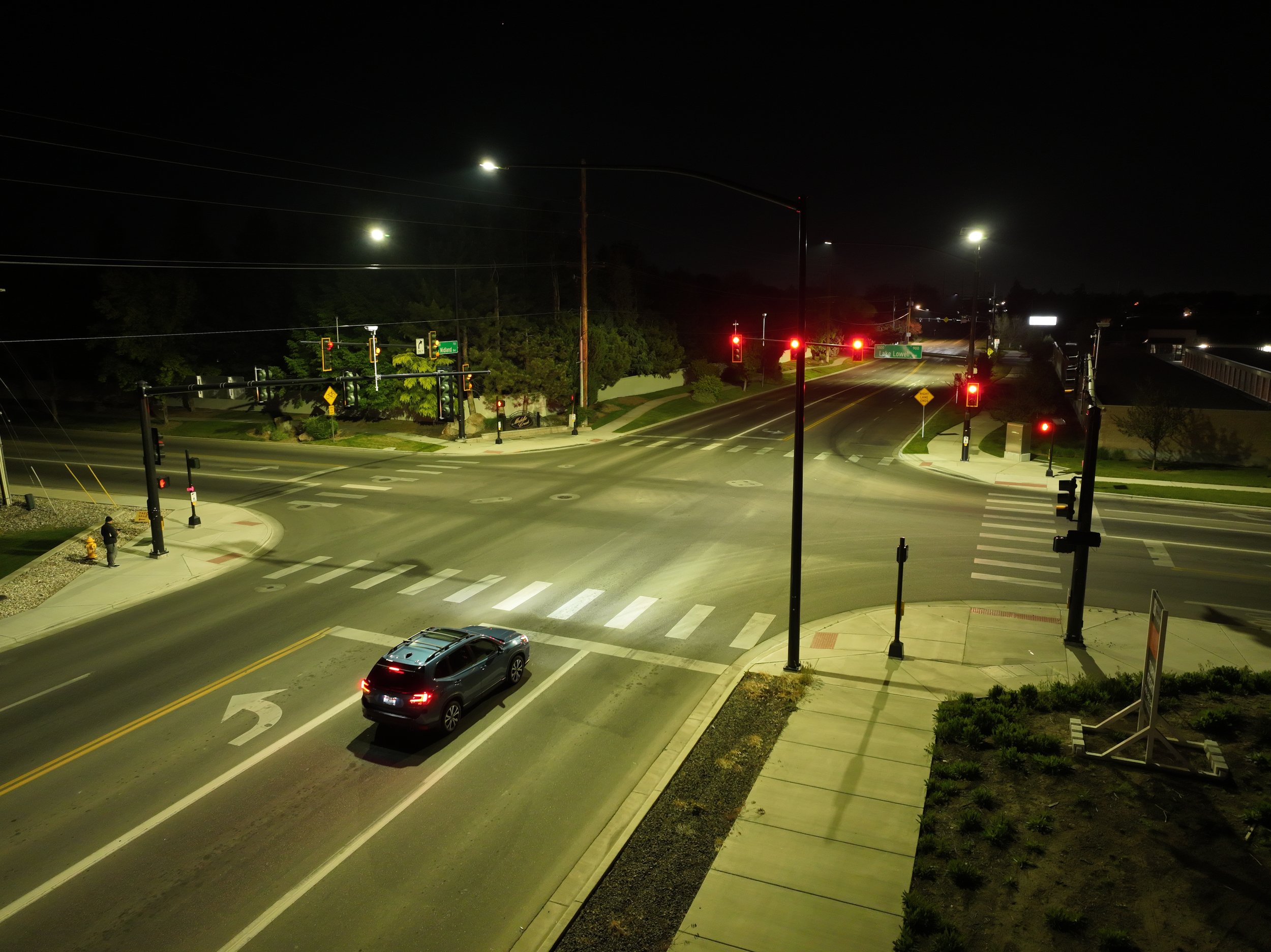 Nighttime view of a city intersection with traffic lights, streetlights, a few cars, trees, and a sidewalk.