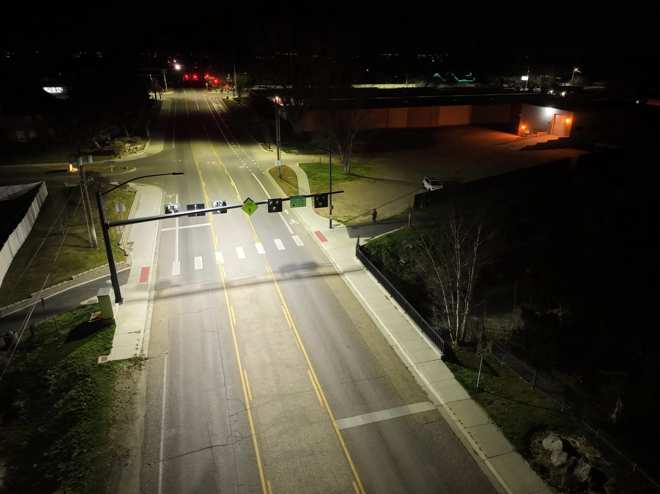 Nighttime view of an intersection with traffic lights, streetlights, and a few parked cars. The streets are empty, and buildings are dimly lit.