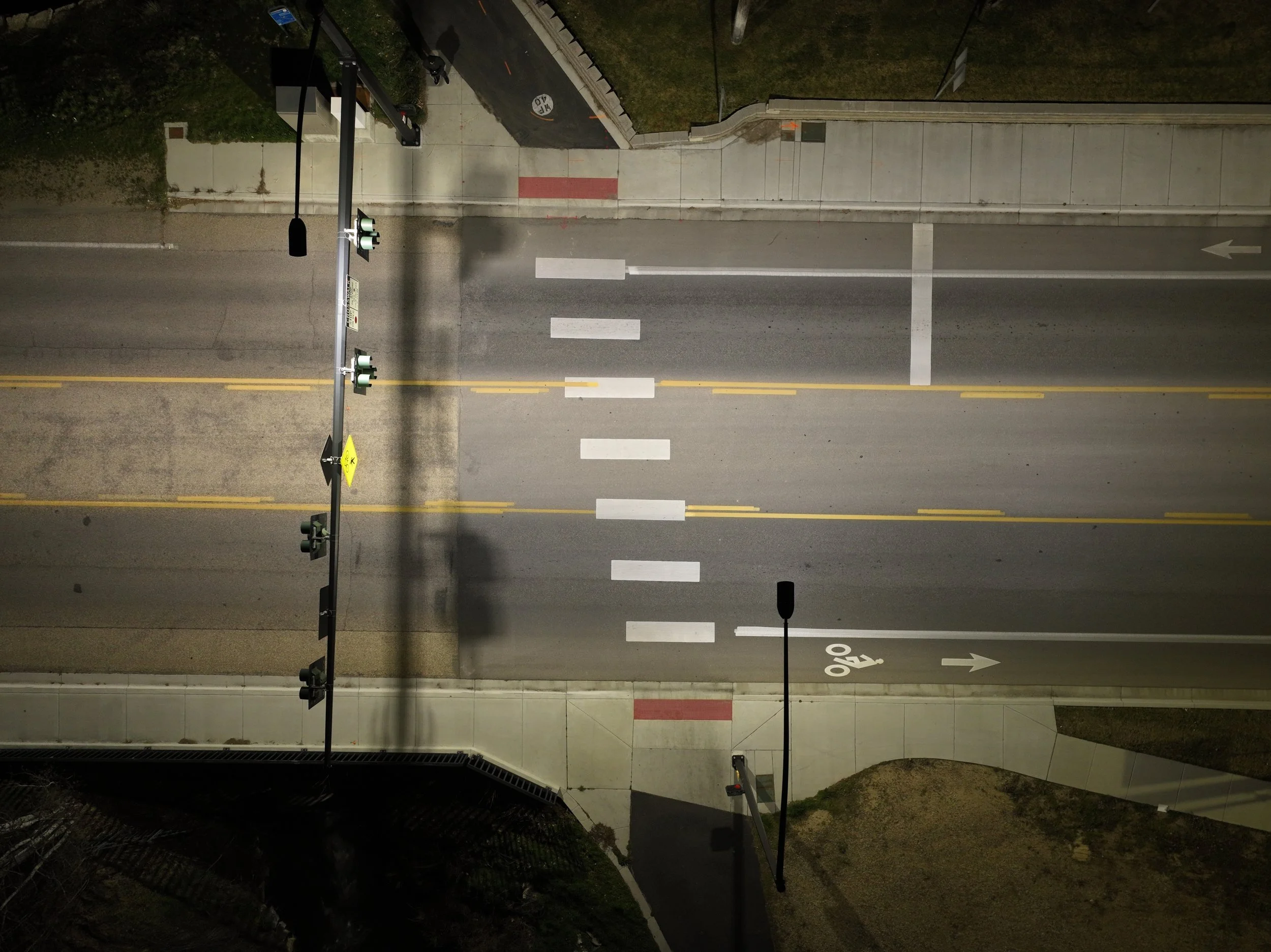A nighttime view of a parking lot with multiple traffic lights, pedestrian crosswalks, and a sidewalk. The parking lot has painted lines for spaces and a bicycle lane. Surrounding areas include grass and building structures.
