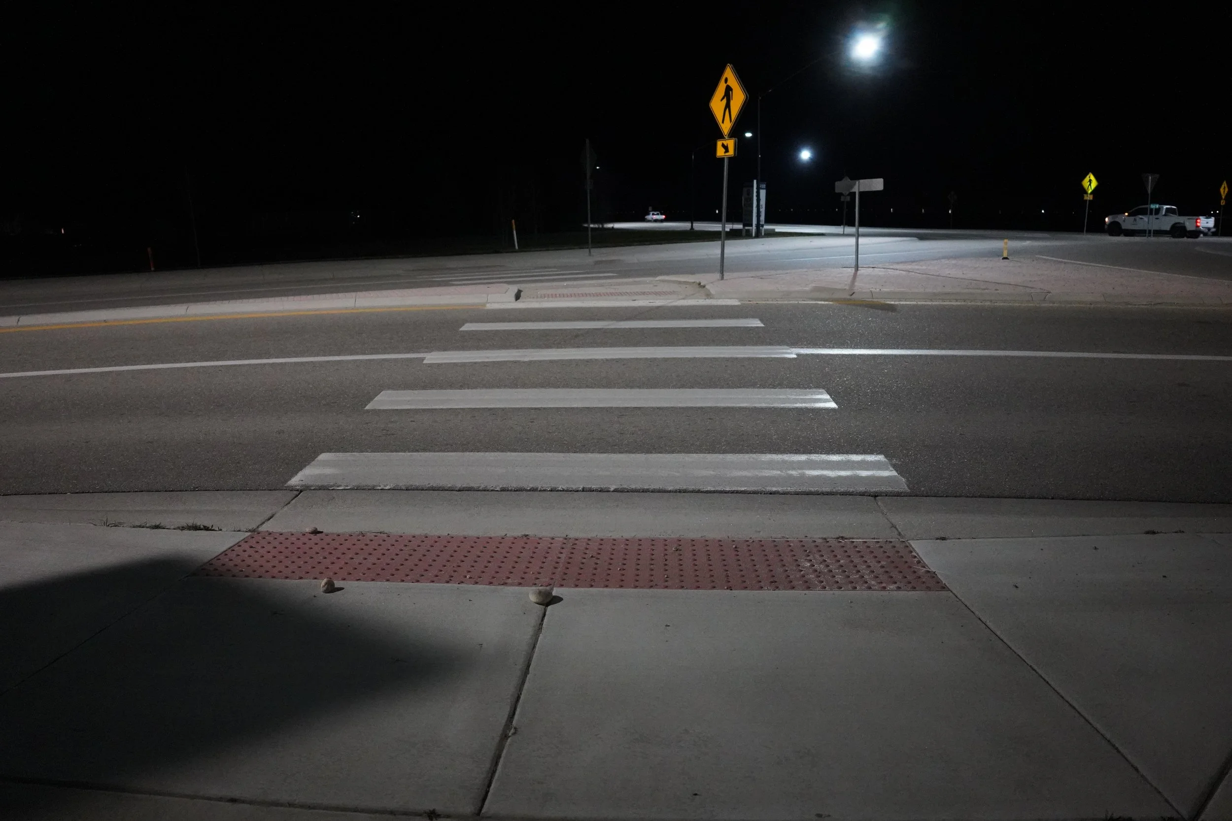 Nighttime view of a crosswalk and pedestrian crossing signs at a street corner, with a pickup truck and a car visible in the distance under the dark sky and bright moon.