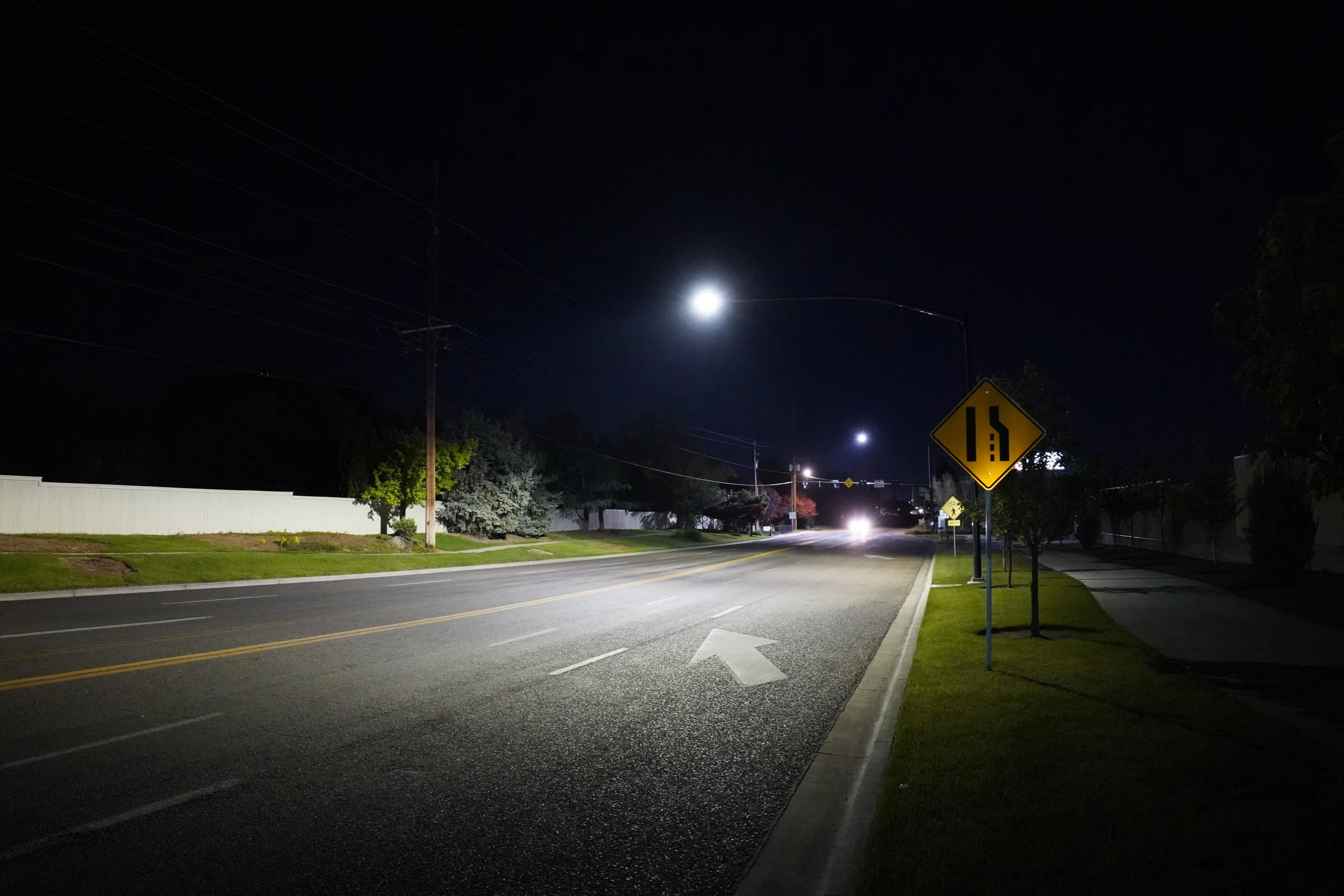 Nighttime street scene showing a dark sky with a bright moon, streetlights, trees, a sidewalk, traffic signs, and an empty road with a white arrow painted on it.