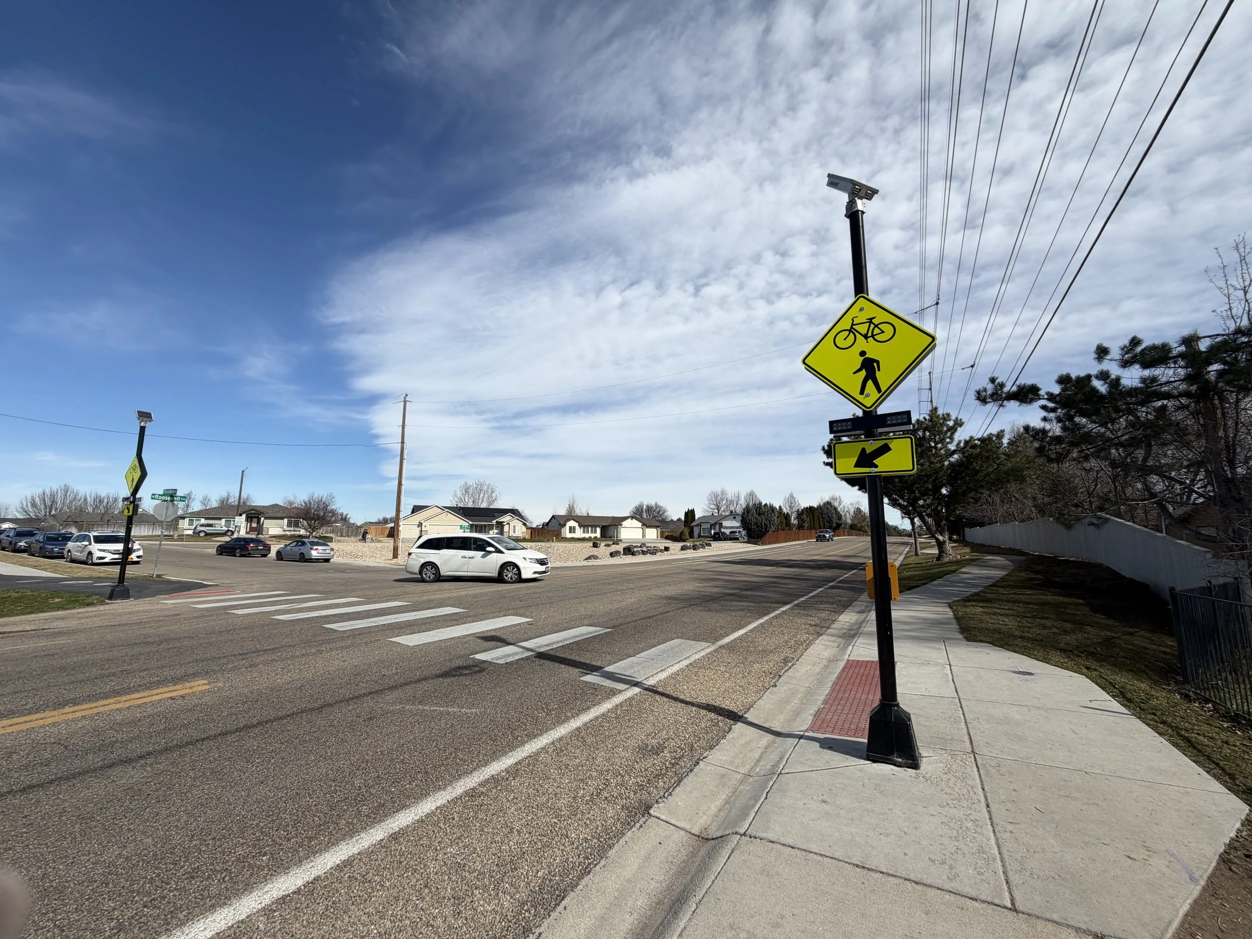 A street corner with a yellow pedestrian and bicycle crossing sign, a traffic light, parked cars, residential houses, and power lines under a partly cloudy sky.