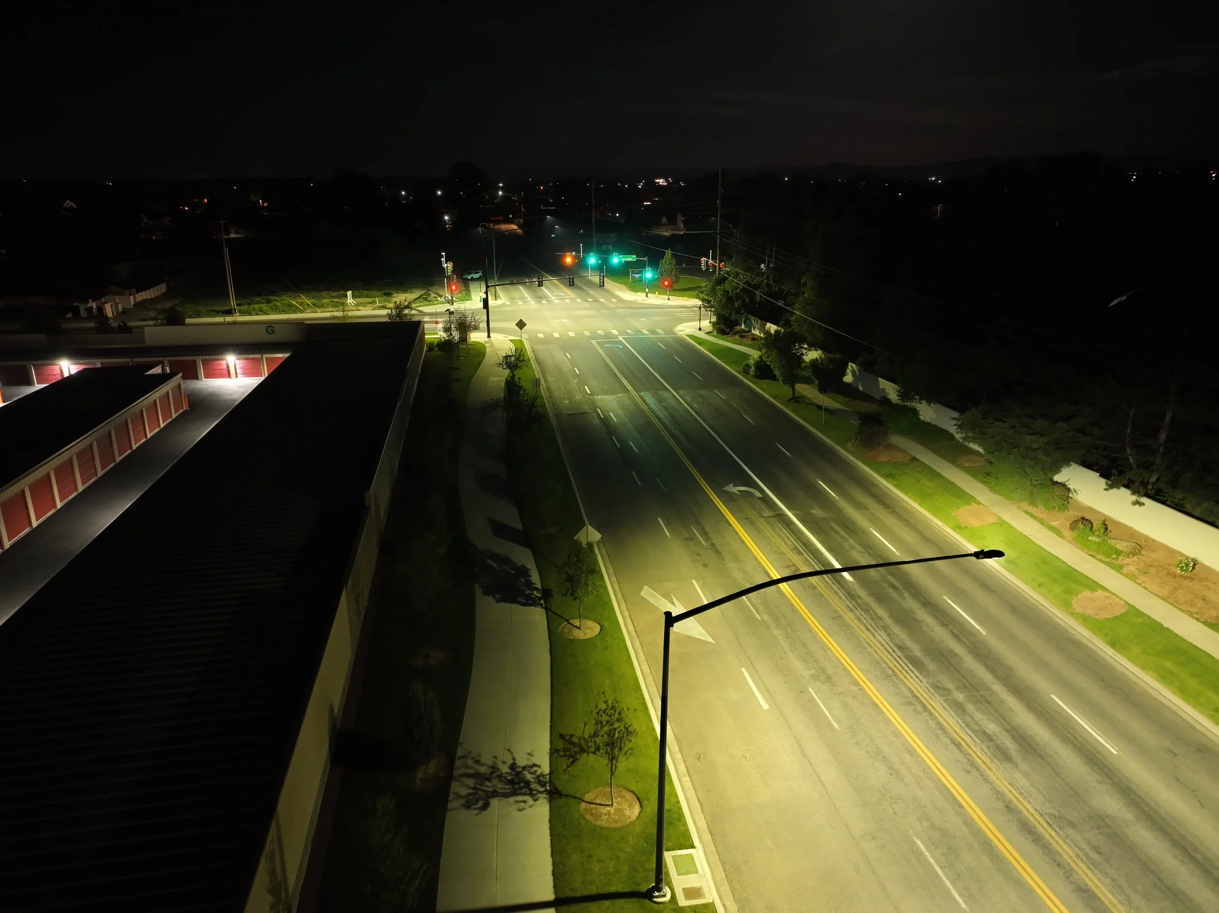 Empty city street at night with green traffic lights, crosswalks, and trees lining the sidewalk. The sky is dark with no visible stars.