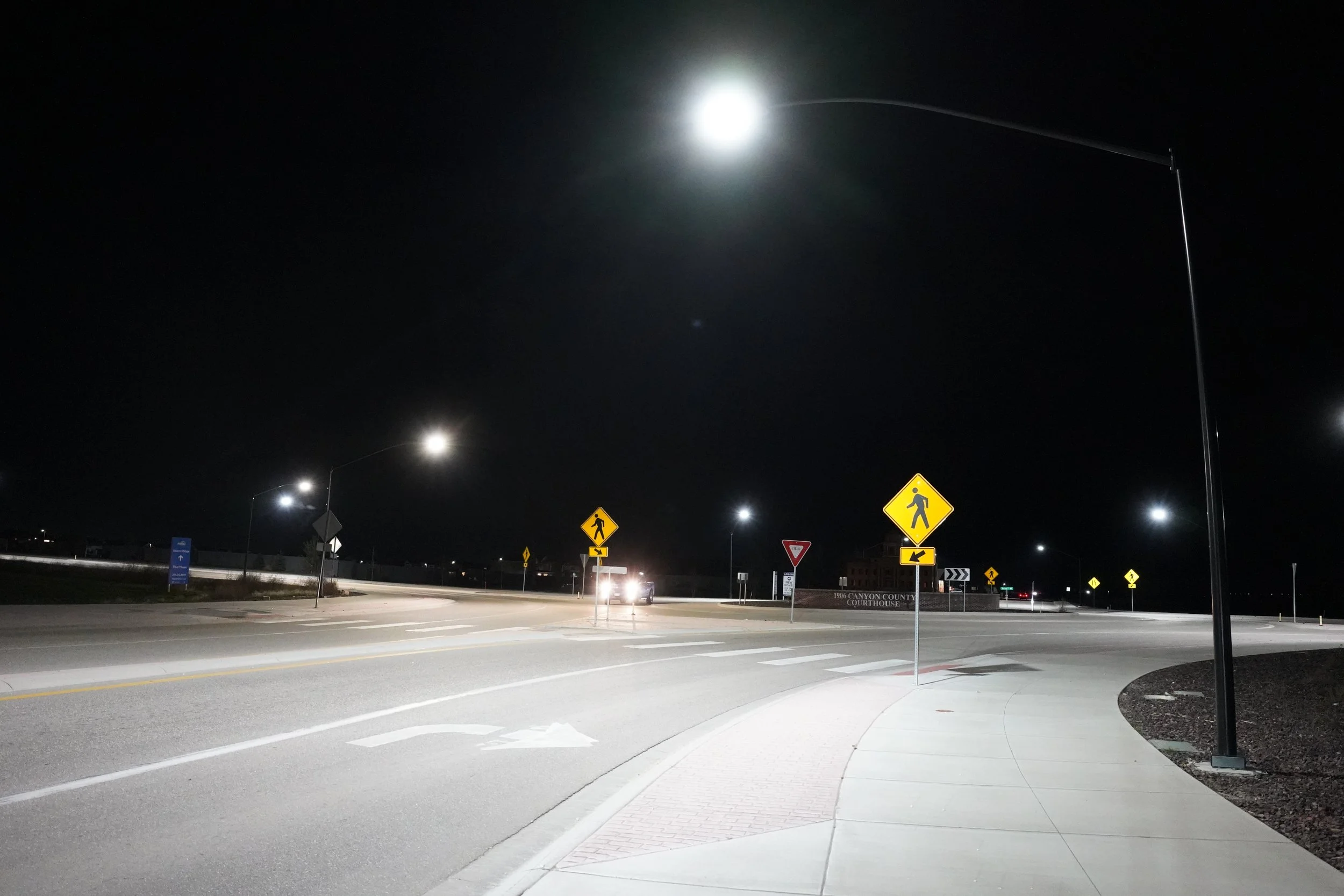 Nighttime view of a city street corner with streetlights and pedestrian crossing signs, including a sidewalk and low-rise buildings in the background.