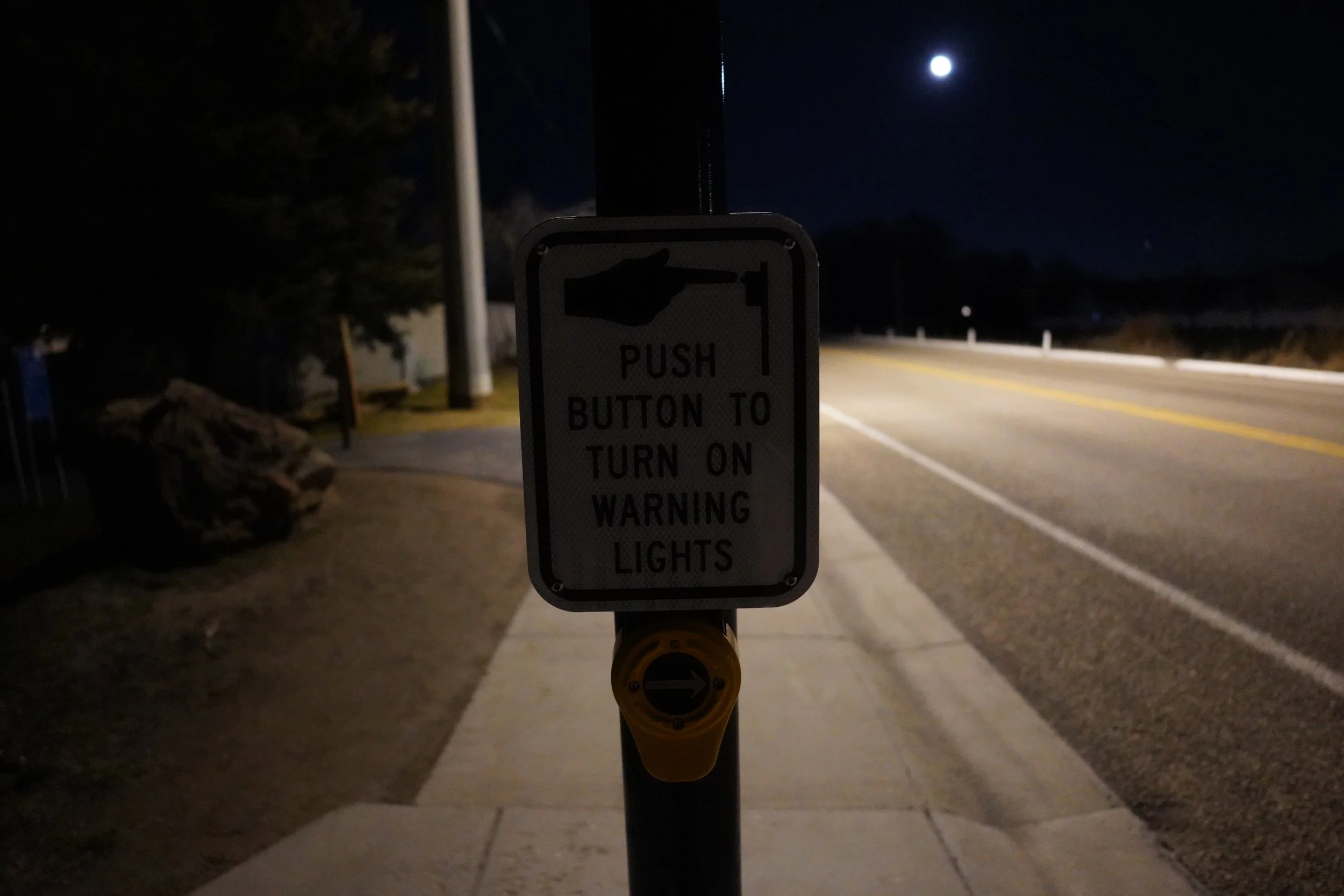 A night scene with a sign on a bicycle or walking path that reads: "Push button to turn on warning lights." The sign has a pointing finger icon above the text. The moon is visible in the sky, illuminating the road and surroundings.