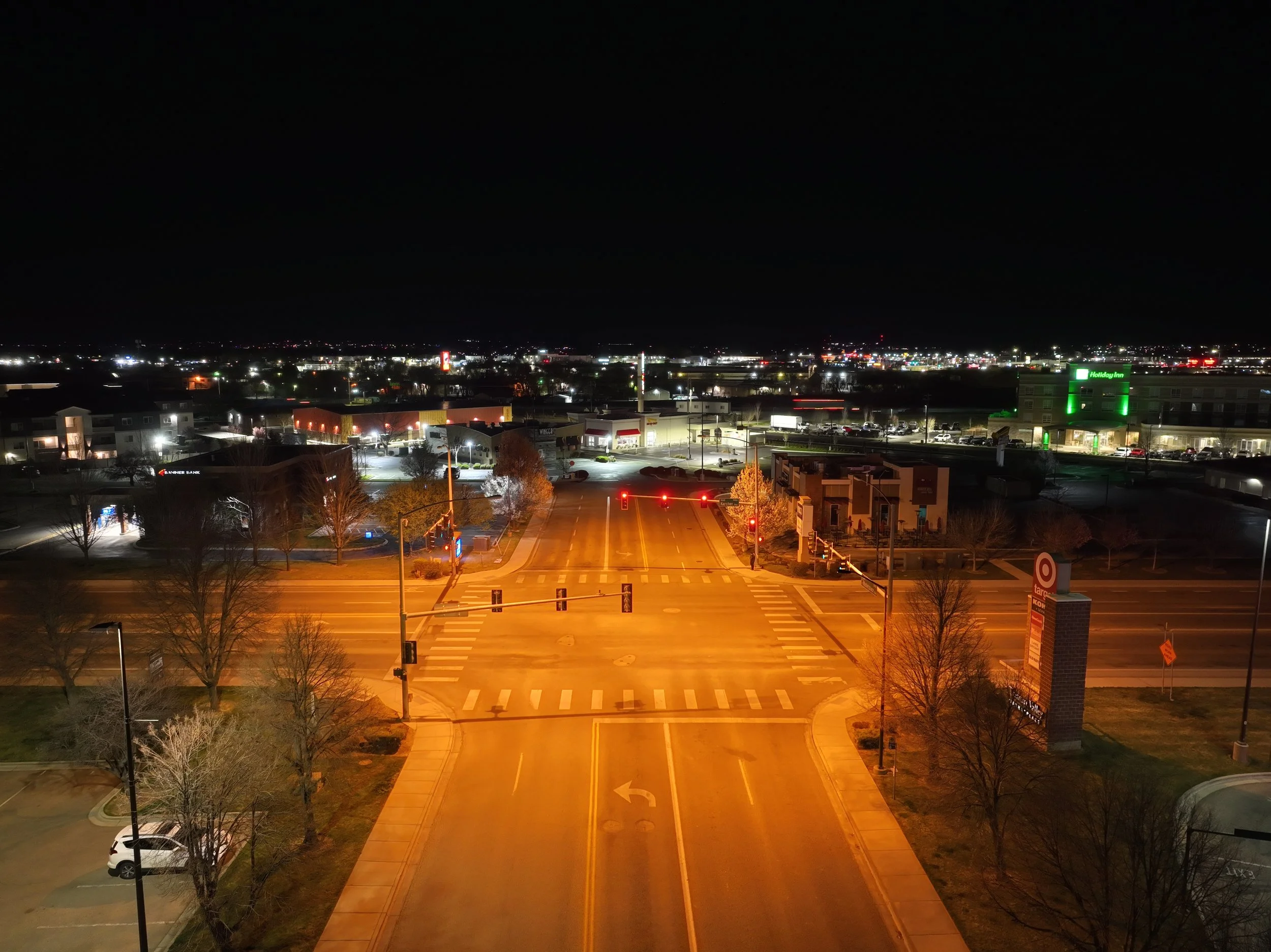Nighttime view of a city with illuminated buildings, streets, and parking lots. There is a large green and white Holiday Inn hotel sign on the right side and traffic lights at the empty intersection in the foreground.