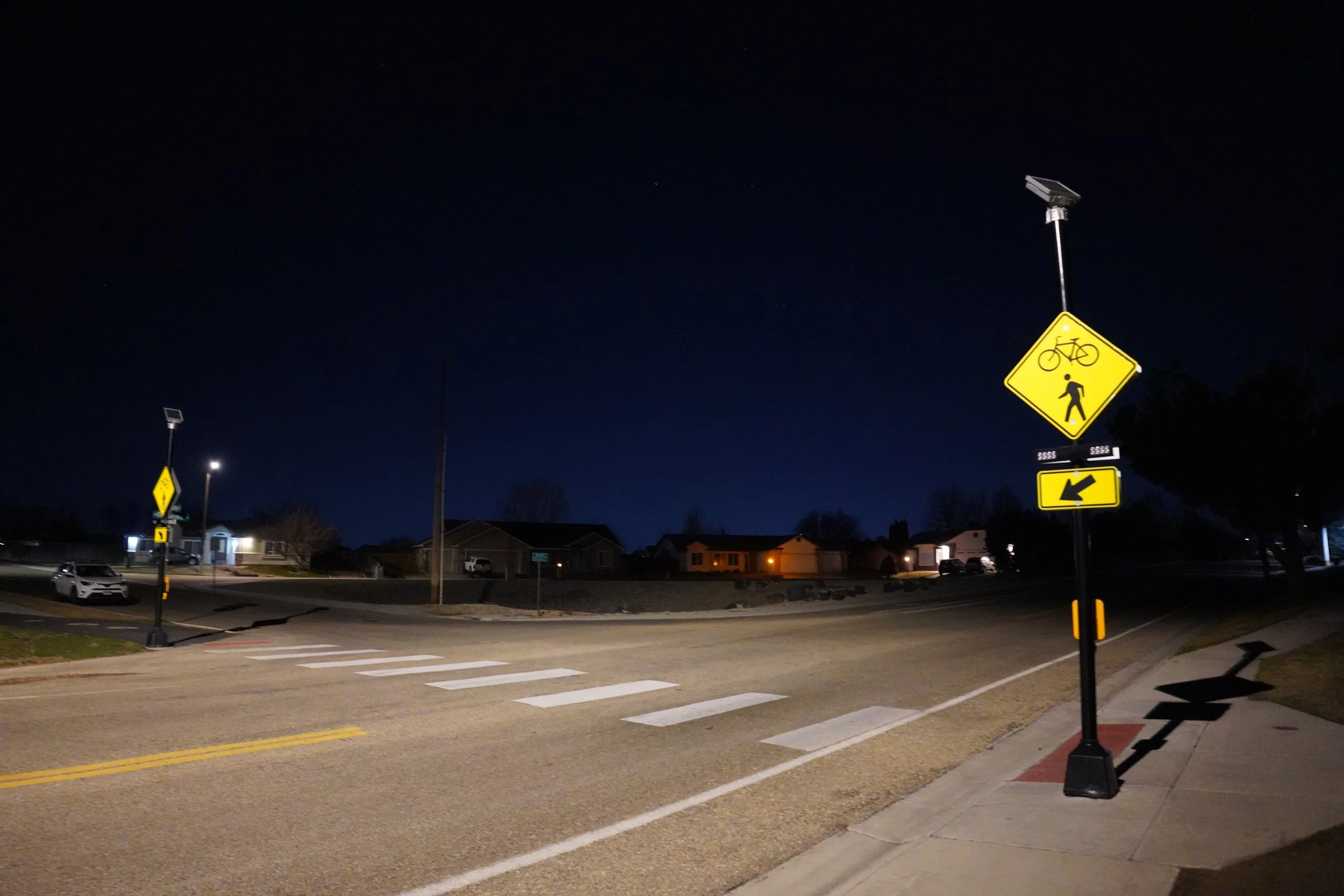 Nighttime view of a street corner with pedestrian and bicycle crossing signs and streetlights illuminating the area. Residential houses can be seen in the background.