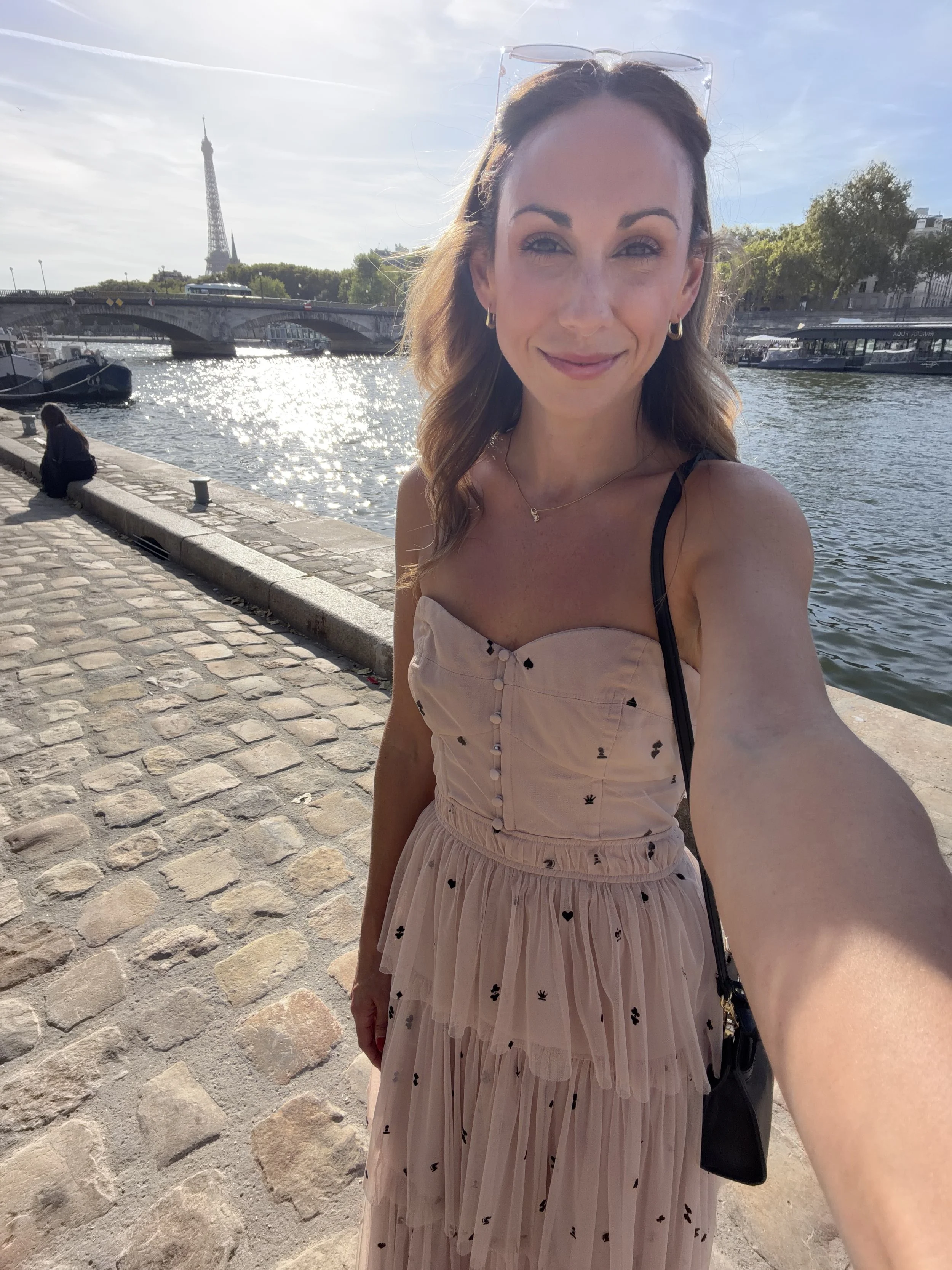 A woman taking a selfie outdoors by the river in Paris with the Eiffel Tower in the background, wearing a light-colored dress with black heart patterns.
