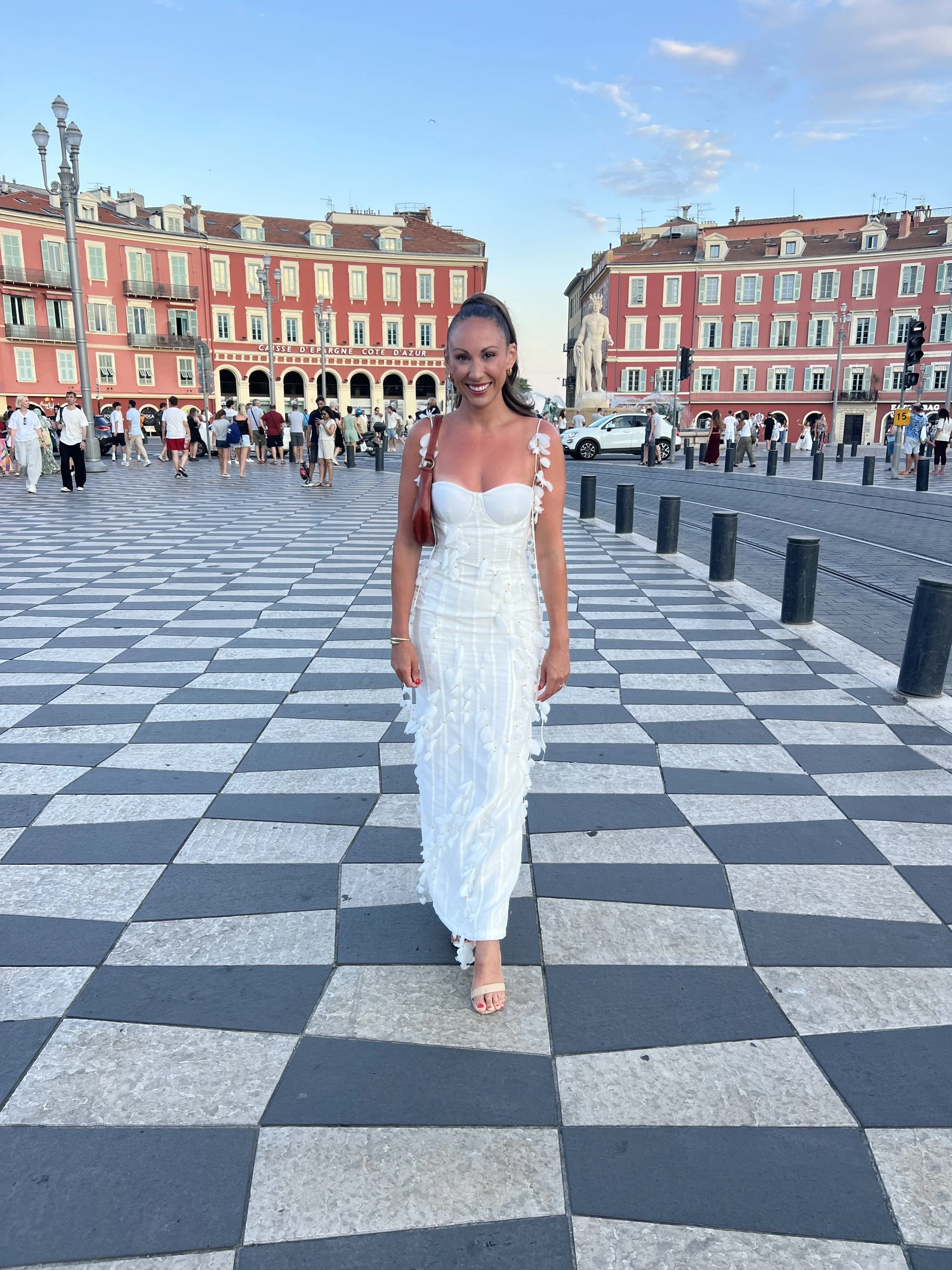 A woman in a white dress with floral details walking on a checkered street in a city square with pink buildings and a statue in the background, during daylight.