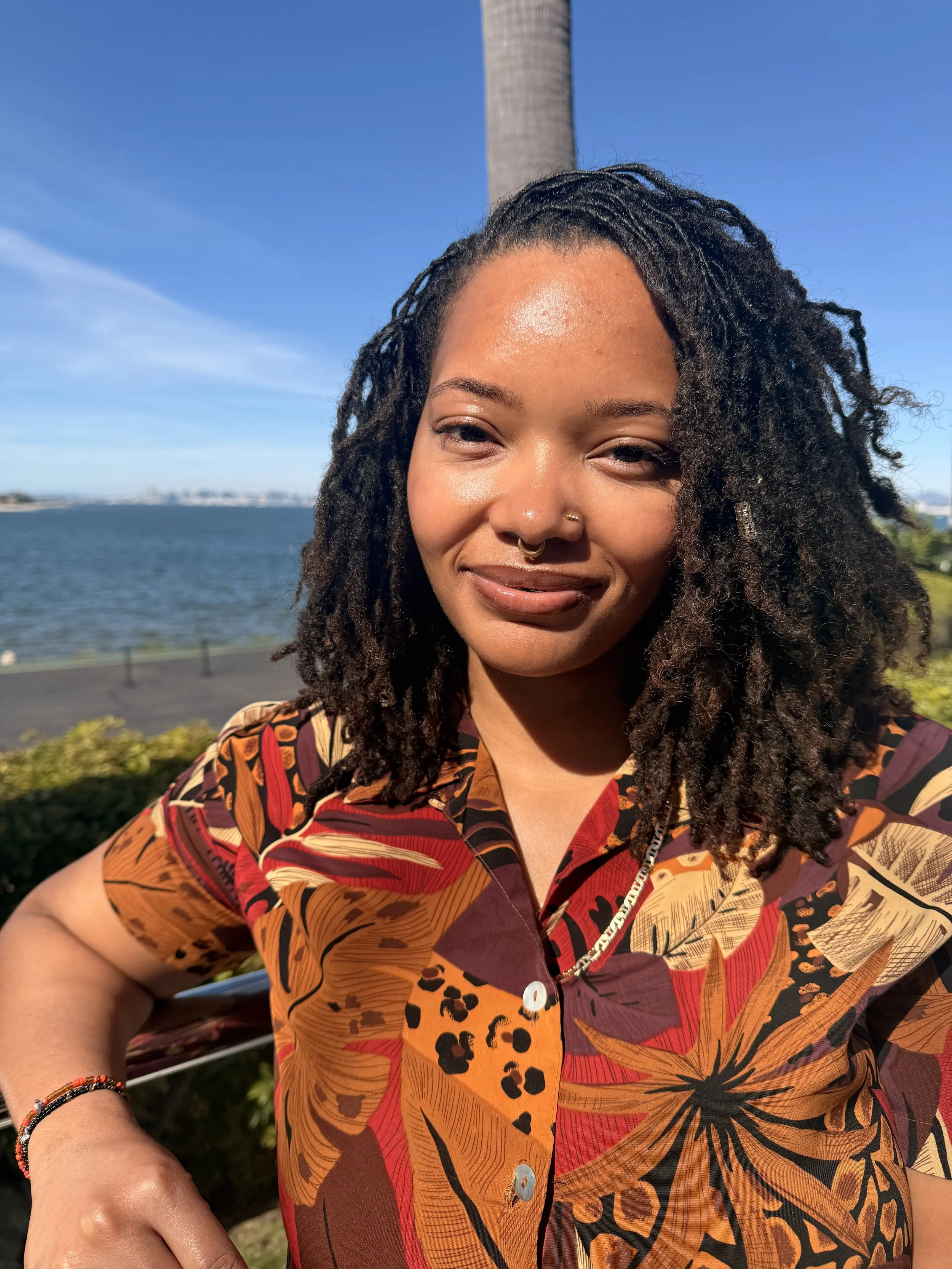 A woman with dreadlocks smiling outdoors near water, with a tall tower and blue sky in the background, wearing a colorful patterned shirt and jewelry.