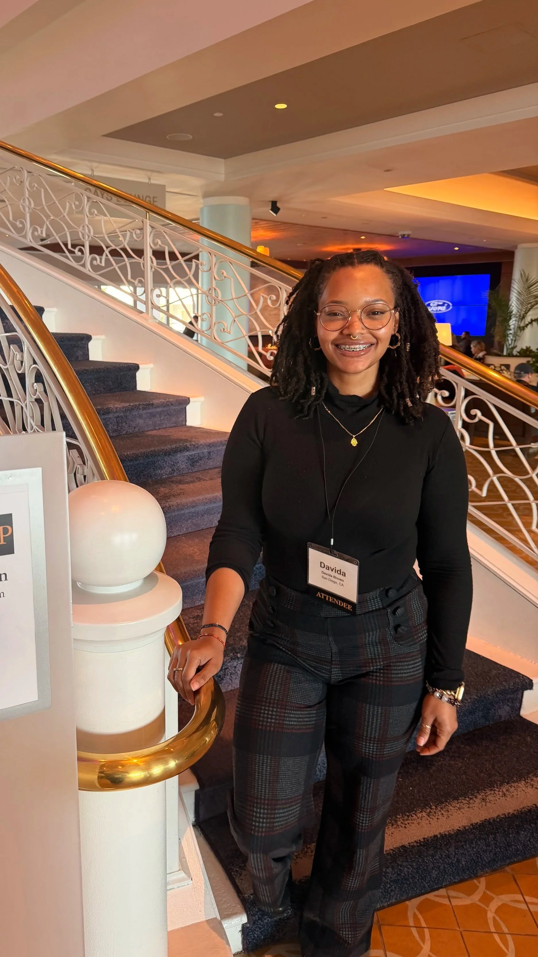 A young woman with dreadlocks, glasses, and a nose ring smiling while standing on a staircase in a hotel lobby. She is wearing a black long-sleeve top, plaid pants, and has a name badge that indicates she is an attendee.
