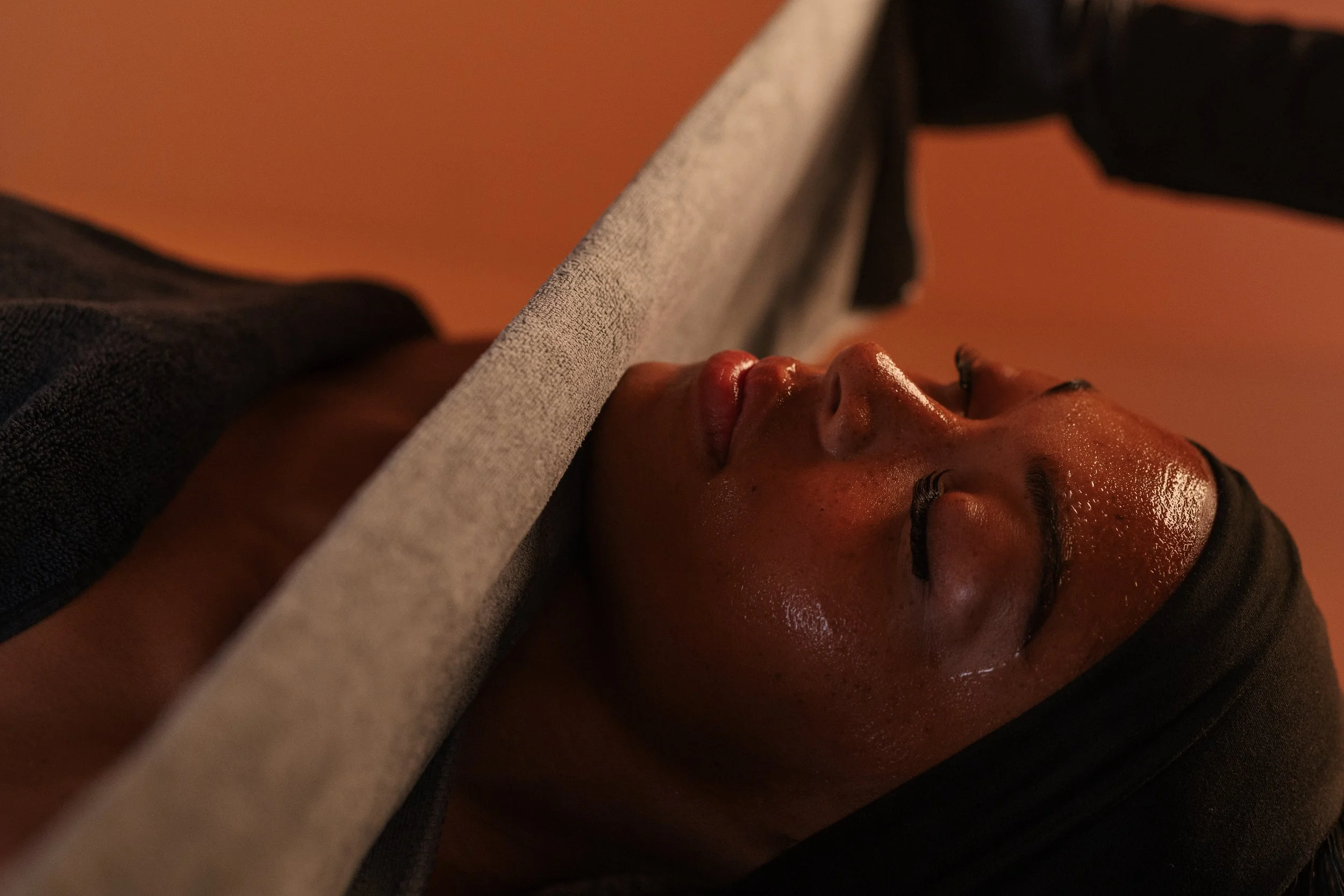 Close-up of a woman with closed eyes relaxing during a massage, with a towel on her forehead and warm lighting in the background.