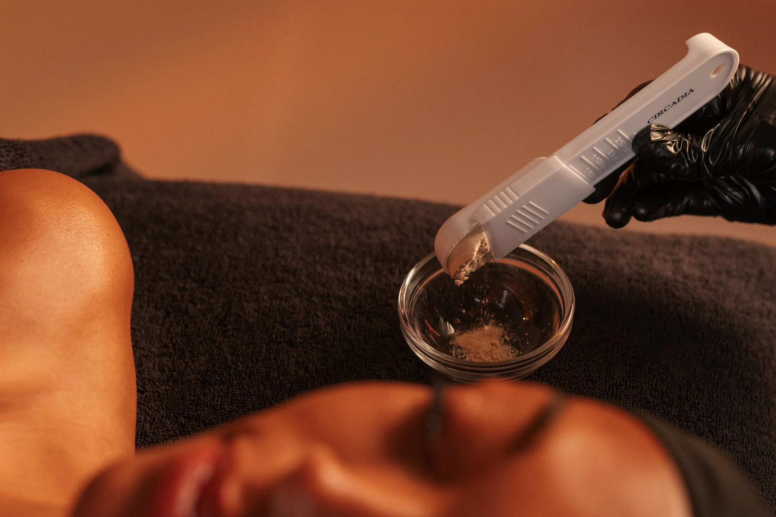 A hand wearing a black glove holds a measuring spoon with active powder over a glass containing a face mask