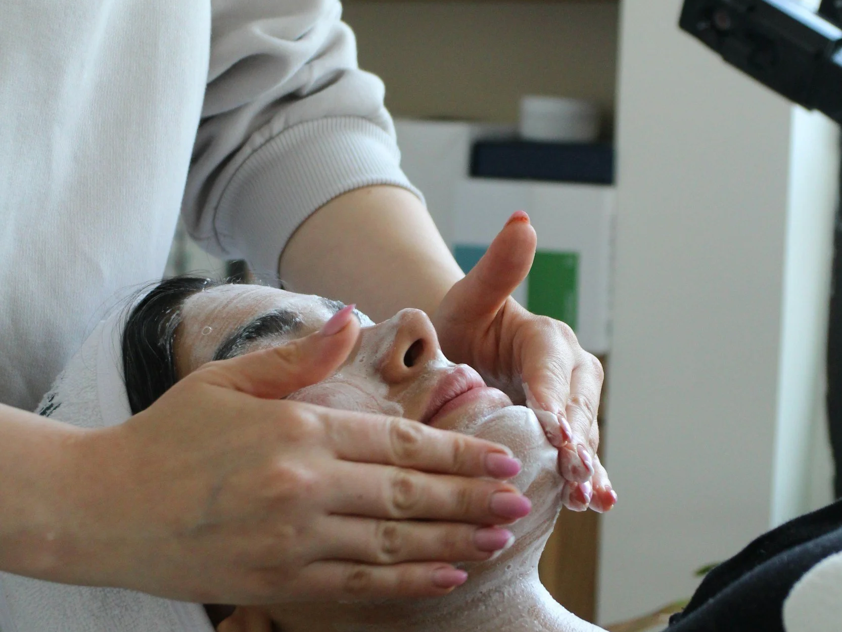 A person receiving a facial treatment, lying down with cleanser on their face, while a practitioner gently massages their forehead and cheeks.