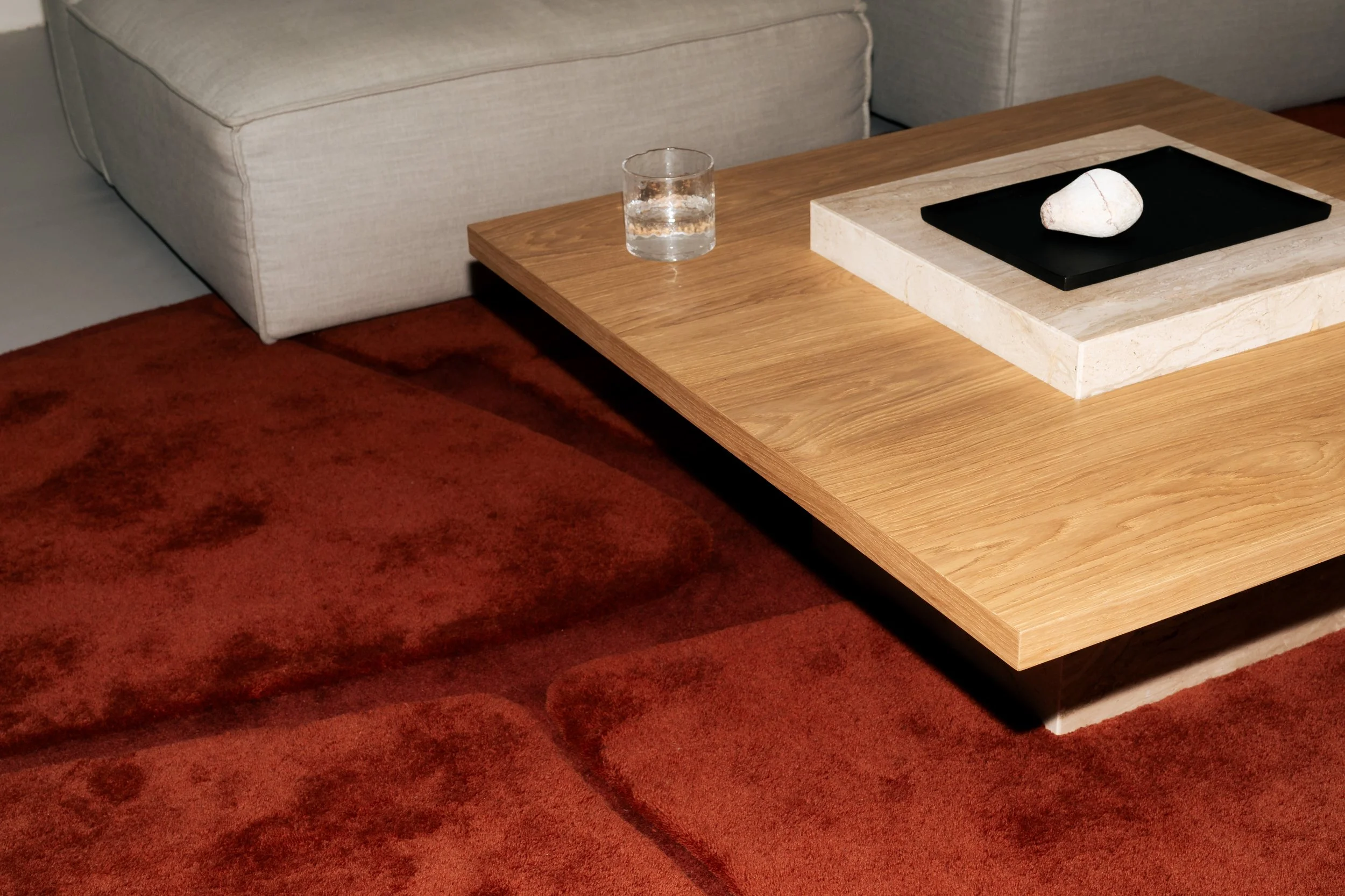 A coffee table with a stone sculpture on top, a glass of water, and a black plate, situated next to a beige sofa and on a red patterned carpet.