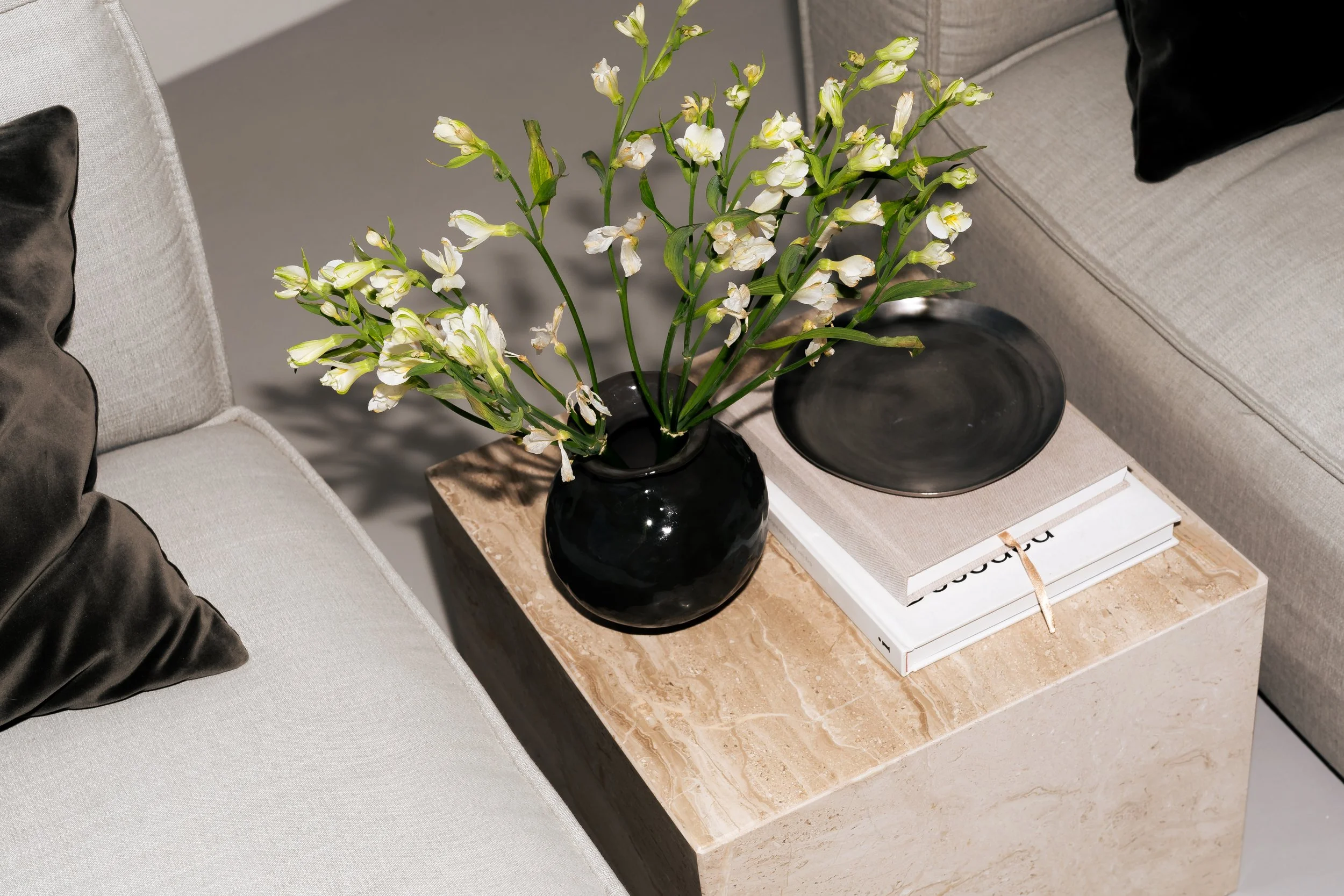 A black vase with white flowers on a beige marble side table next to a gray couch with black pillows.