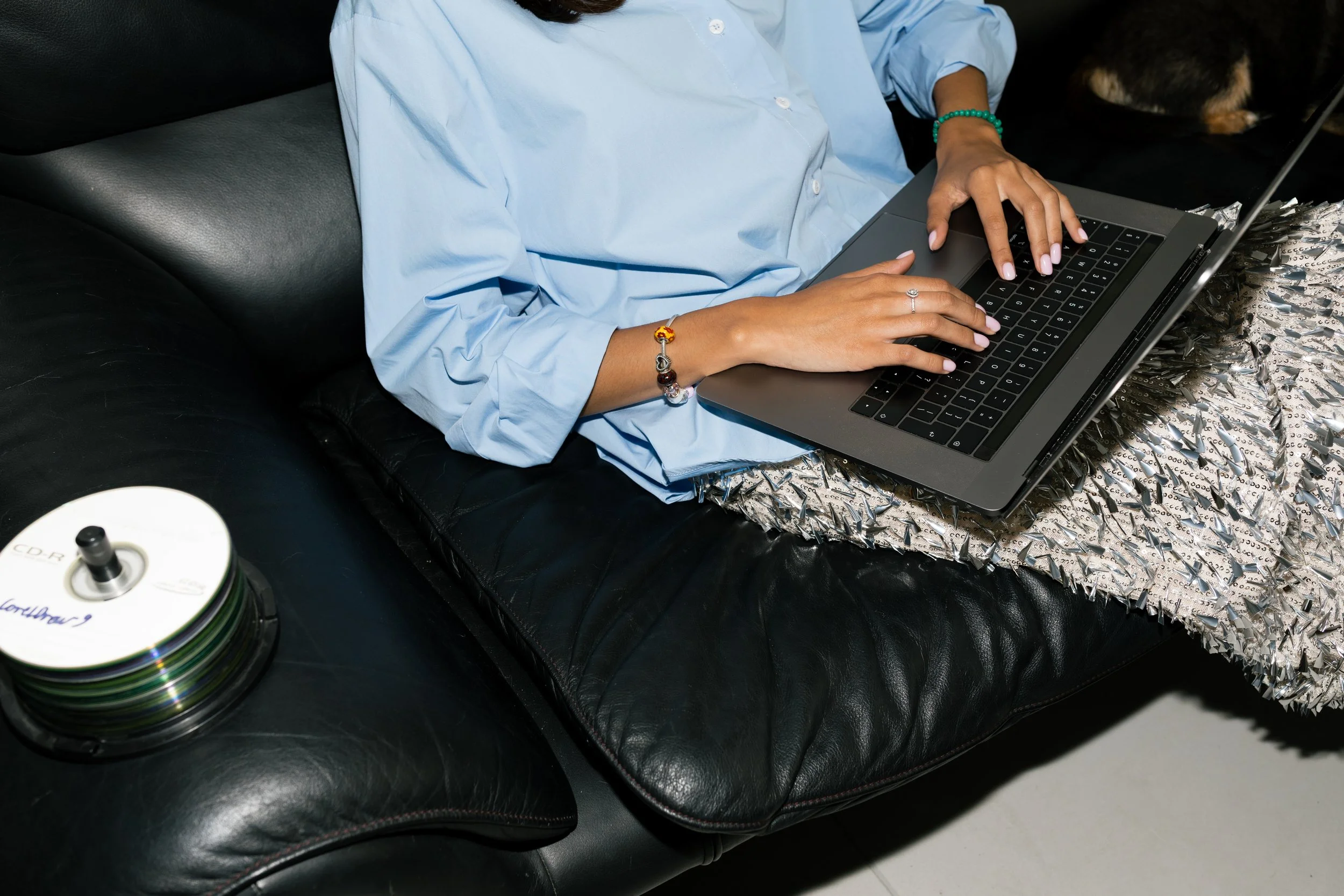 Person wearing a light blue shirt and jewelry, using a MacBook laptop on a textured surface, on a black leather couch, with a stack of CDs nearby.