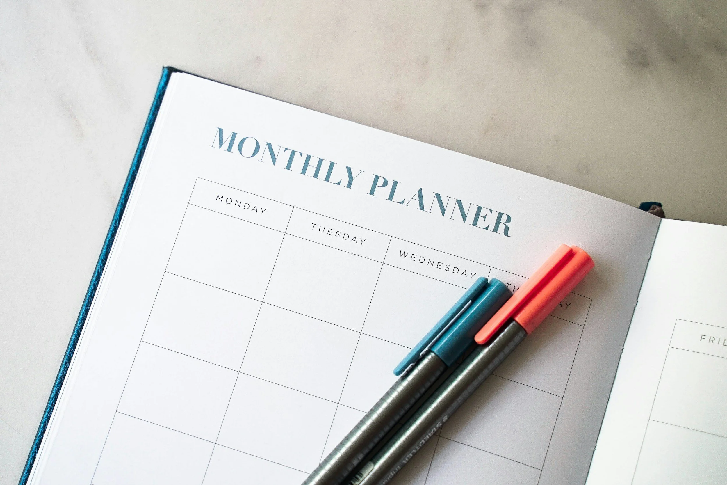Open monthly planner with days of the week visible, accompanied by several markers in blue, gray, and coral colors resting on the pages.