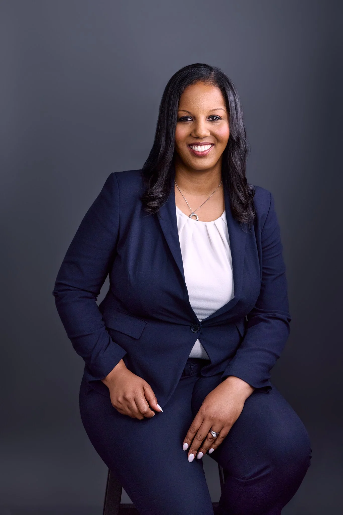A woman with black hair, wearing a navy blazer and white top, sitting against a dark gray background, smiling with hands resting on her lap.