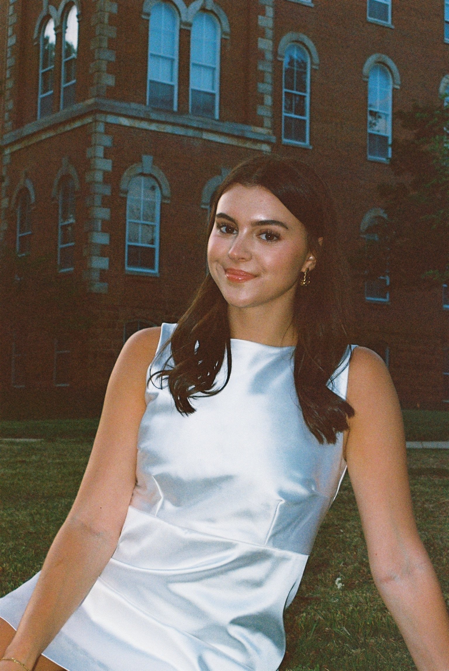A young woman with long, dark hair and light skin, wearing a white sleeveless dress, is smiling and sitting on the grass in front of a brick building with multiple arched windows.