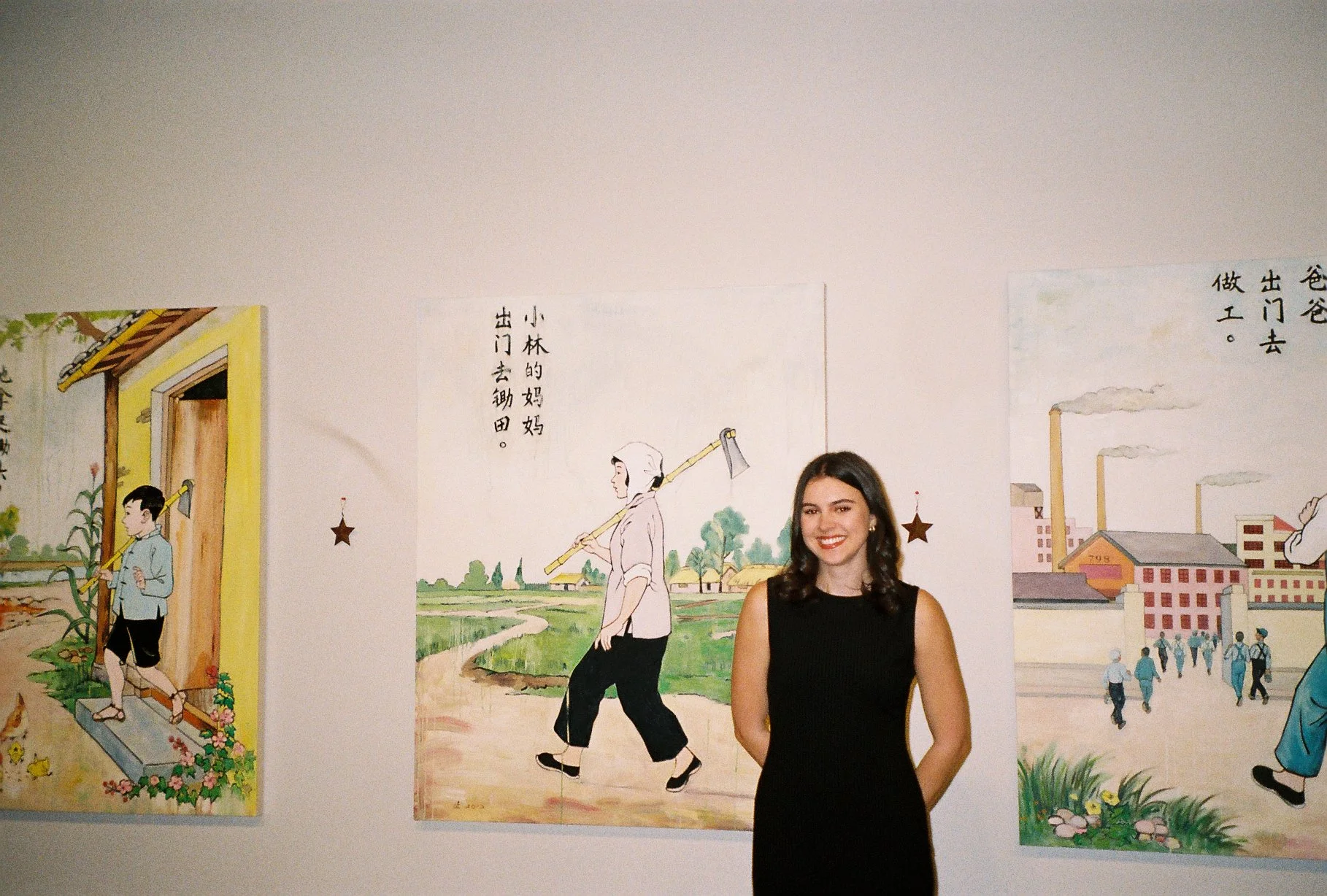 A young woman in a black sleeveless dress standing in front of colorful illustrations on a wall, depicting scenes with people and buildings with Chinese characters next to them.