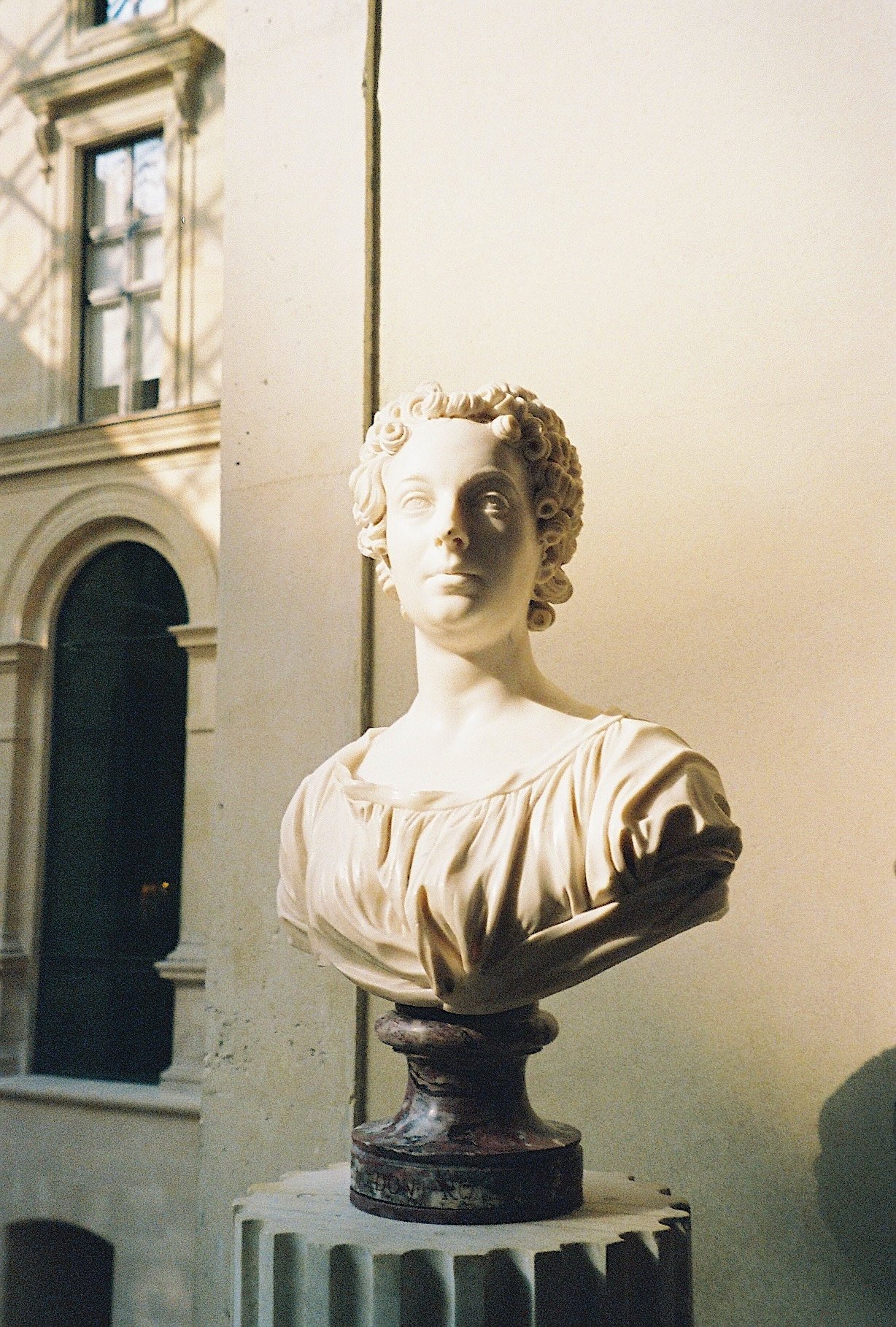 A marble bust sculpture of a woman with curly hair, dressed in a pleated garment, displayed on a dark pedestal.