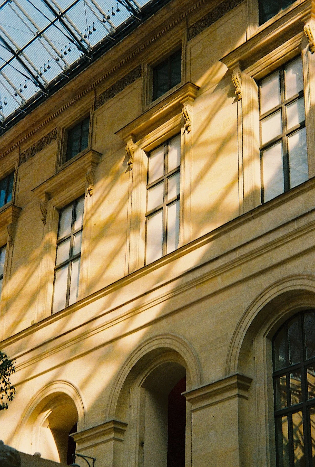 A historic building with ornate architectural details, large arched windows, and decorative moldings. Sunlight casts shadows across the facade, and a glass ceiling or canopy is visible at the top of the image.