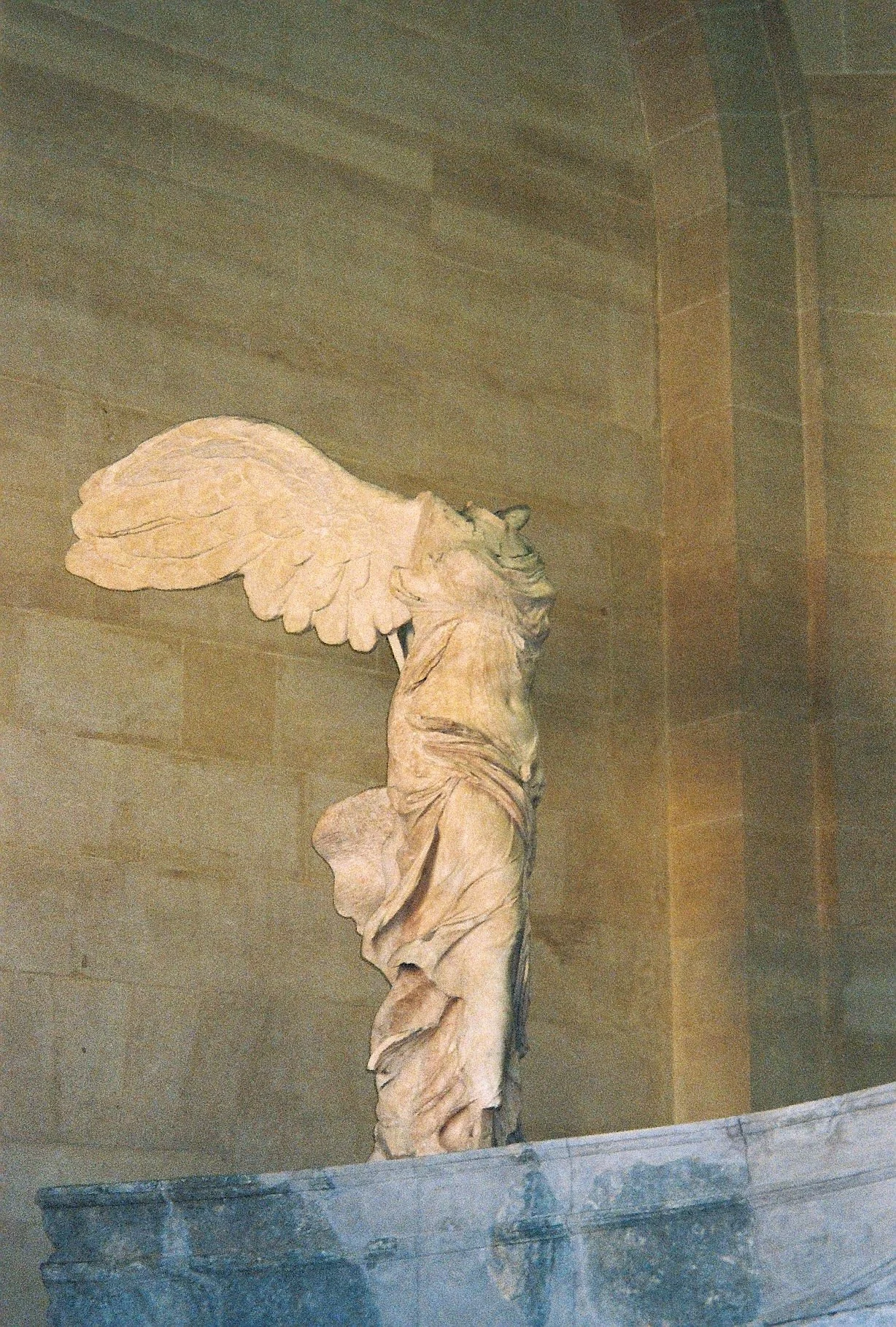 A classical marble sculpture of a winged figure, possibly a mythological or angelic being, displayed on a stone pedestal against a beige stone wall.