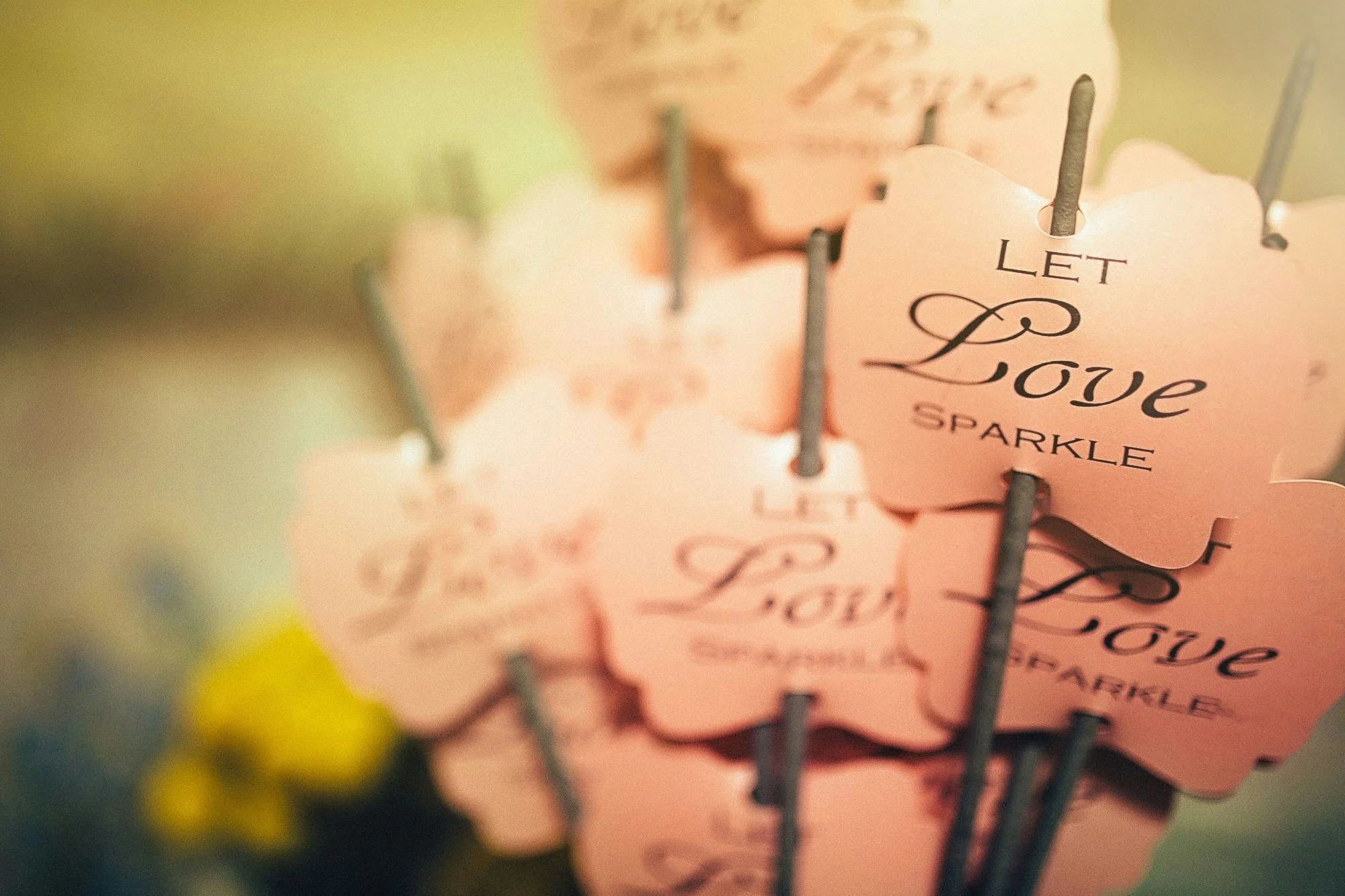 Close-up of pink heart-shaped tags with the words 'Let Love Sparkle' written in cursive, attached to silver sticks with a blurred background.