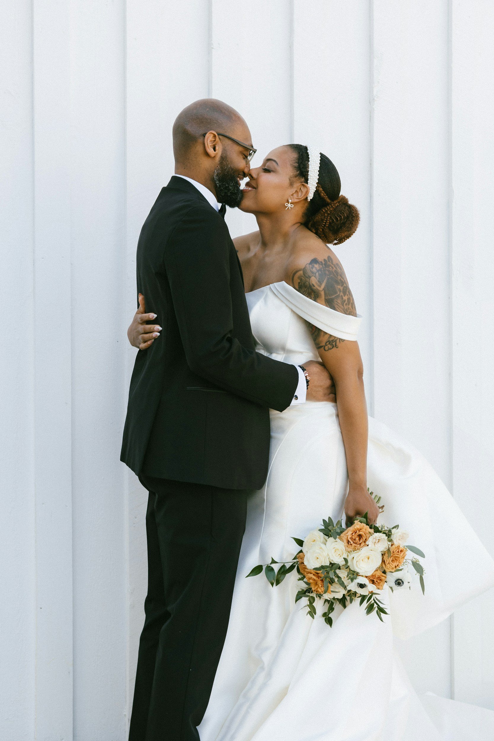 A bride and groom stand close together, smiling and touching noses, in front of a white wall. The bride has tattoos on her right arm, wears a pearl headband and off-the-shoulder wedding dress, and holds a bouquet of white and blush flowers with green foliage. The groom wears a black tuxedo, glasses, and has a beard and a shaved head.