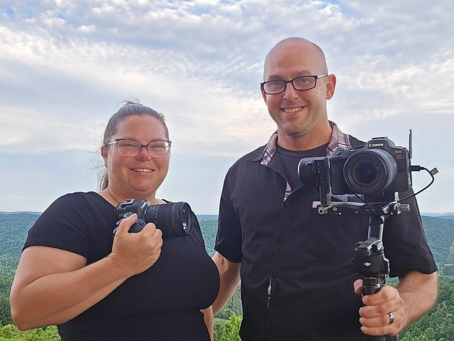 A smiling man and woman videographers standing outdoors, both holding professional cameras, with scenic landscape and cloudy sky in the background.