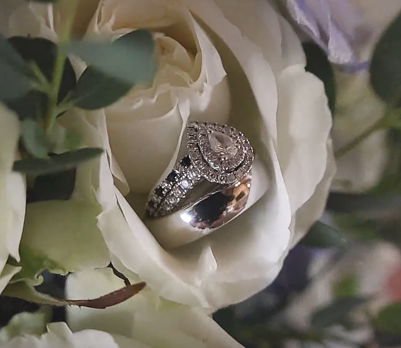 Close-up of a silver ring with diamonds, placed inside a white flower with green leaves surrounding it for a wedding