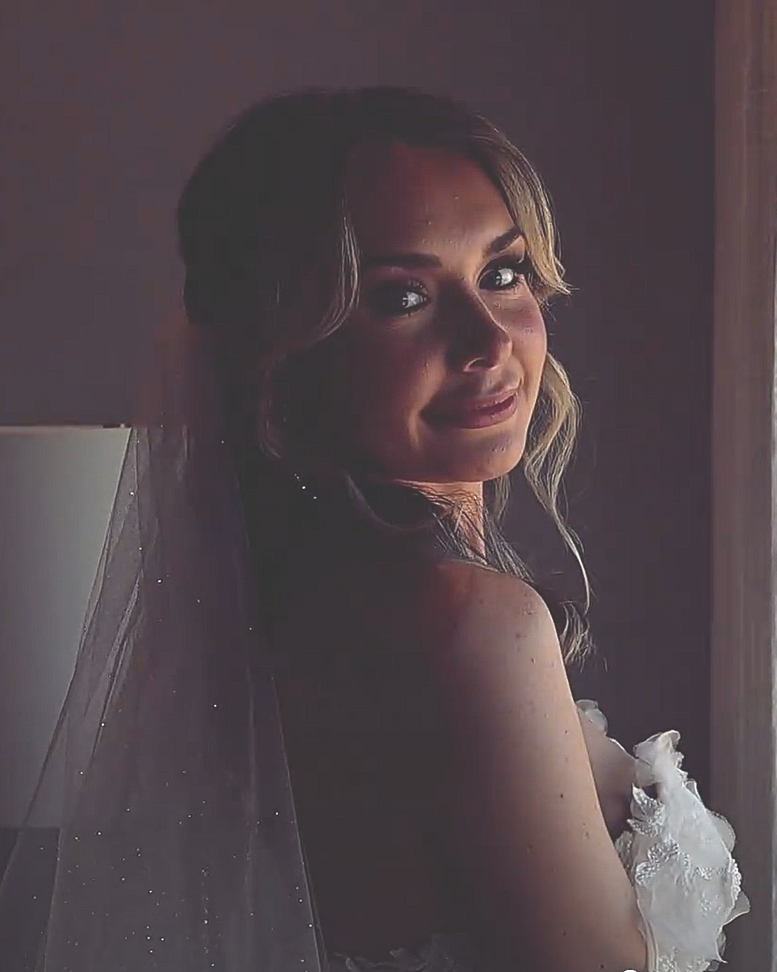 A woman with light brown hair styled in loose waves, wearing a strapless white dress with lace details, looking over her shoulder with a gentle smile, standing near a window with soft lighting.