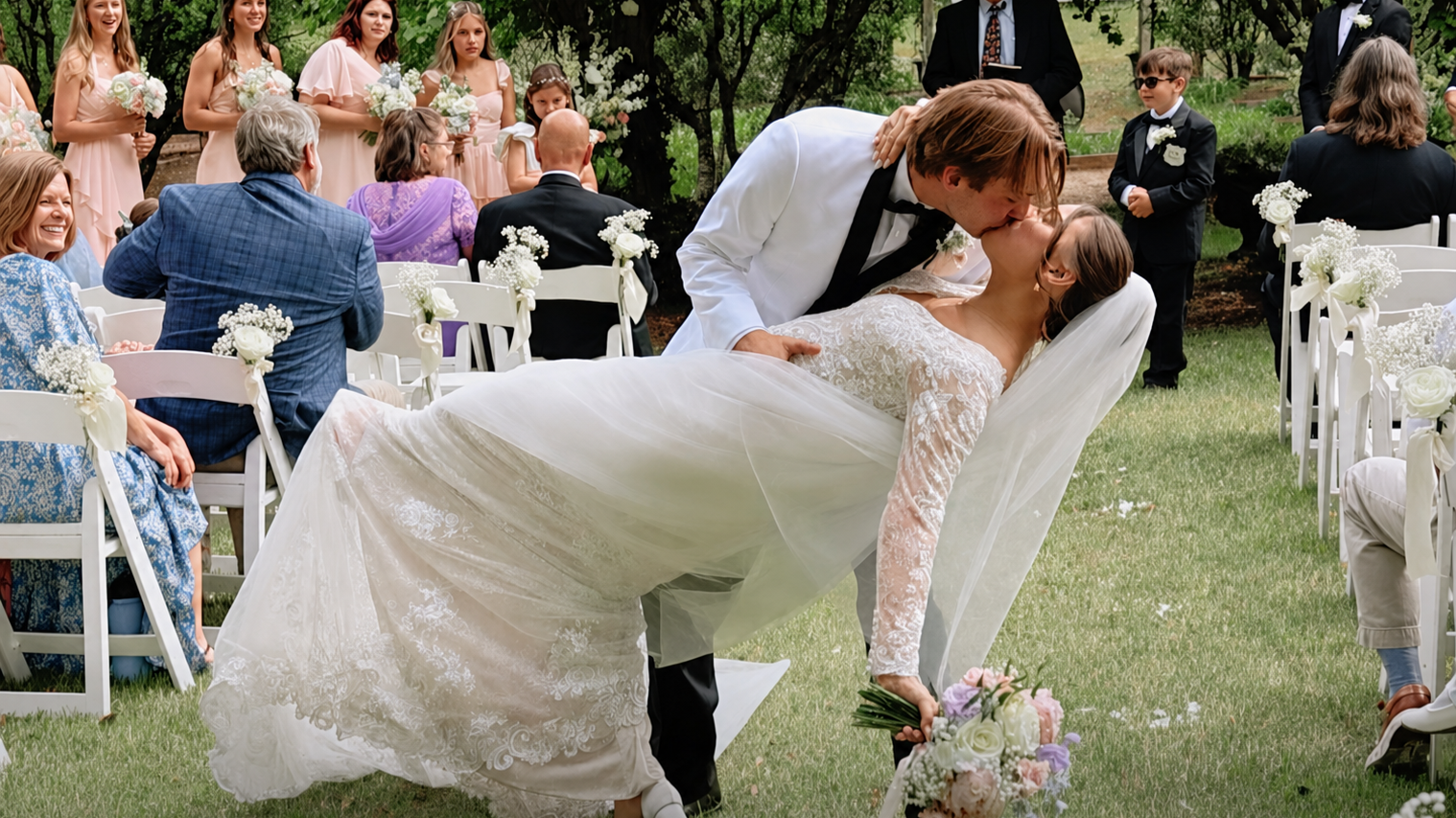 A wedding ceremony outdoor with a bride and groom kissing as the groom dips her. The bride holds a bouquet of flowers. Guests are seated on white chairs decorated with flowers, and bridesmaids in pink dresses stand in the background.