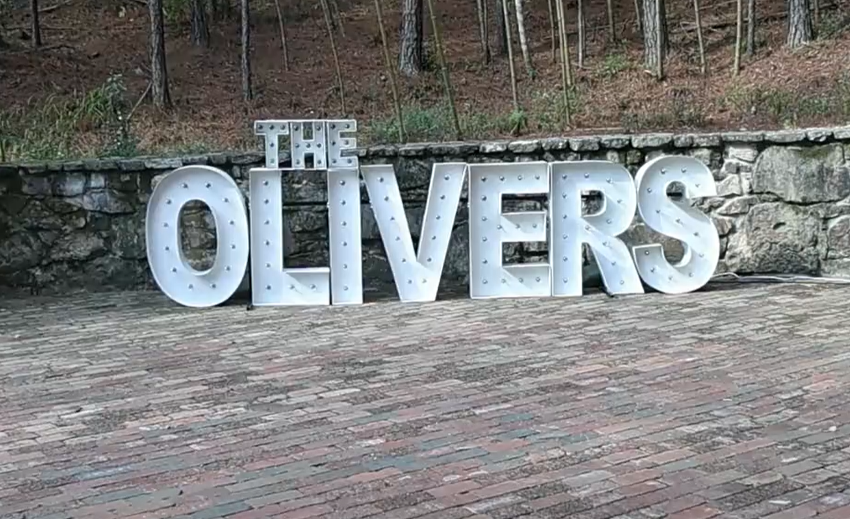 Large white illuminated marquee letters spelling 'THE OLIVERS' placed on a cobblestone surface with a stone wall and trees in the background.