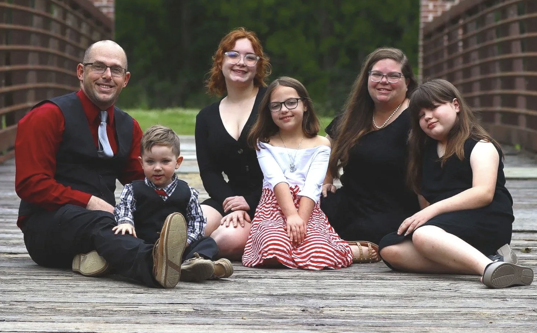 Family of six sitting on a wooden dock outdoors with green trees and a brick structure in the background during a sons adoption.