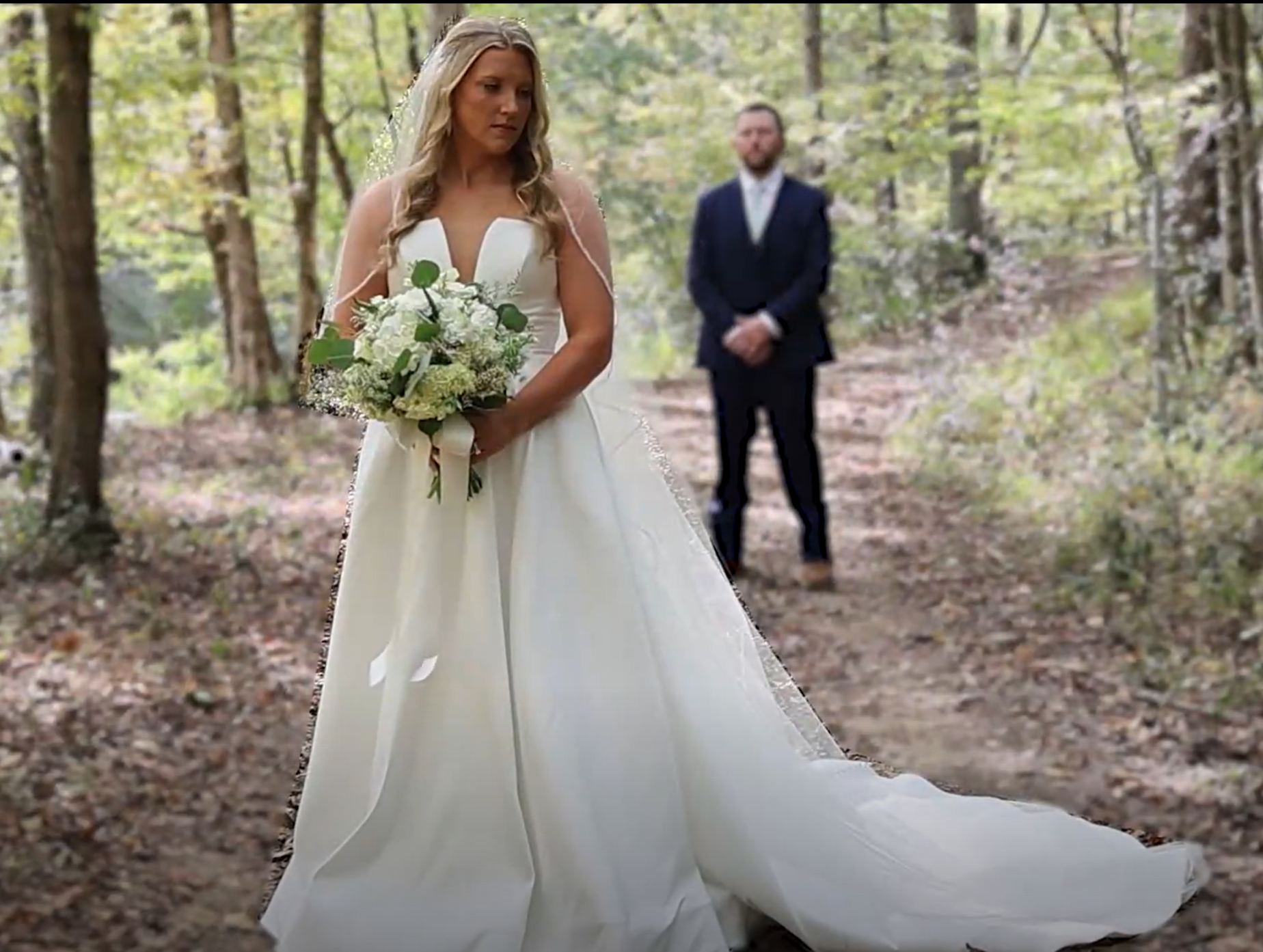 A bride in a white wedding gown holding a bouquet of white flowers stands in a wooded outdoor setting, with a groom dressed in a dark suit standing behind her in the background.