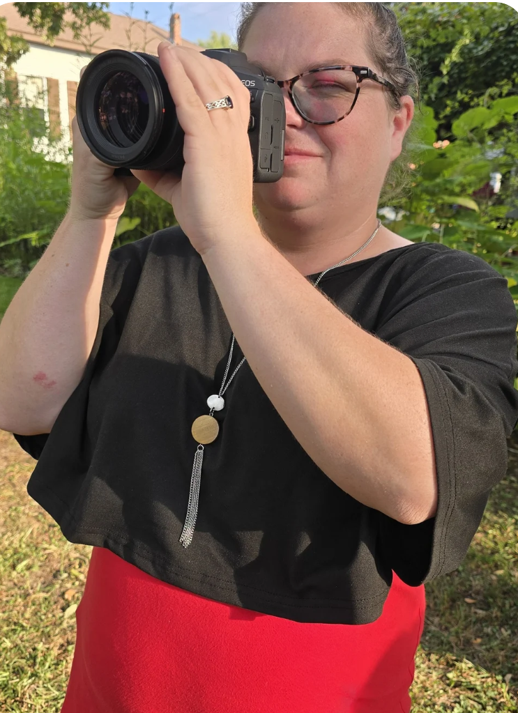 Woman with glasses and a necklace taking a photograph outdoors during daytime, standing in a garden with green foliage and a house in the background.