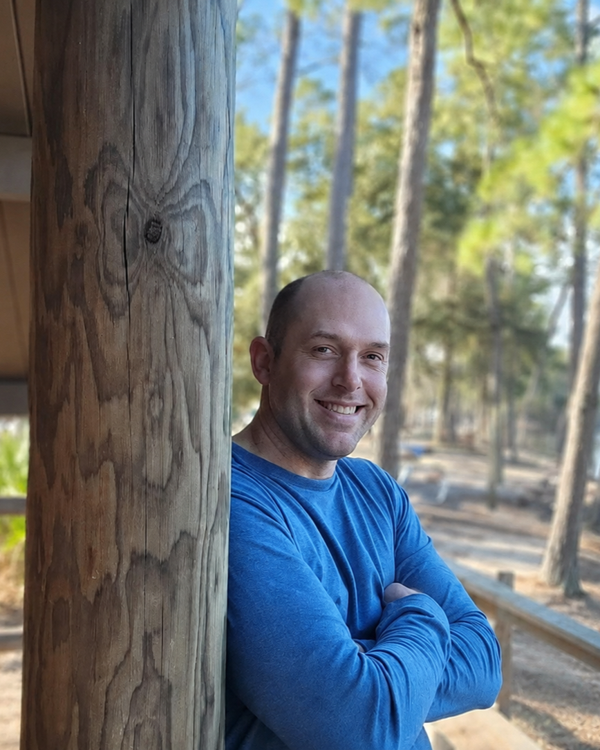 A man in a blue long-sleeve shirt leaning against a wooden post outdoors, smiling with arms crossed, with trees and a path in the background.