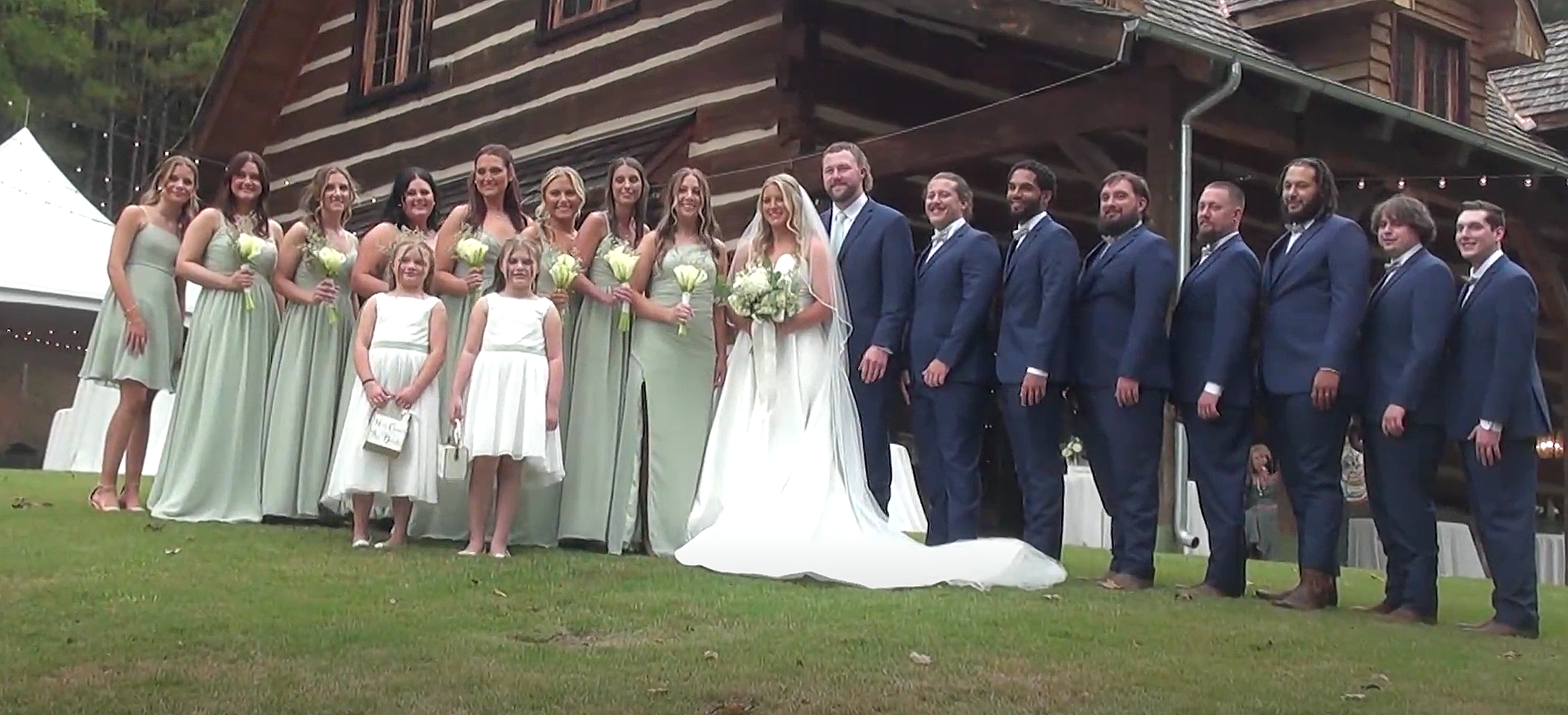 A wedding party standing outside a rustic wooden building, with women in light green dresses holding bouquets, men in navy blue suits, a groom standing beside a bride, and a bride in white holding a bouquet, all smiling.