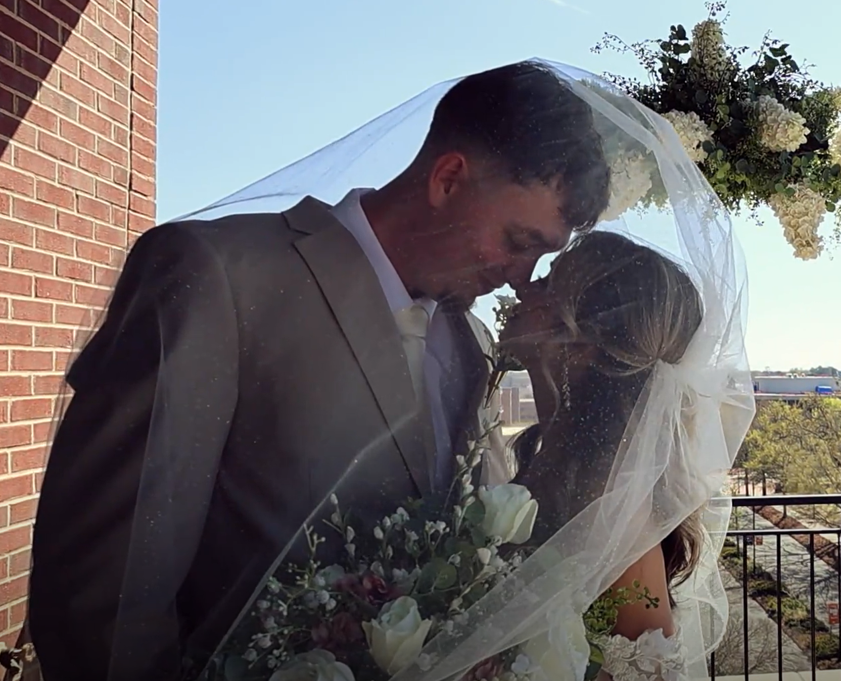 A bride and groom sharing a kiss at their wedding, with a bouquet of flowers visible. The bride has a veil, and the groom is wearing a suit. They are outdoors next to a brick wall and a metal railing, with trees and a blue sky in the background.