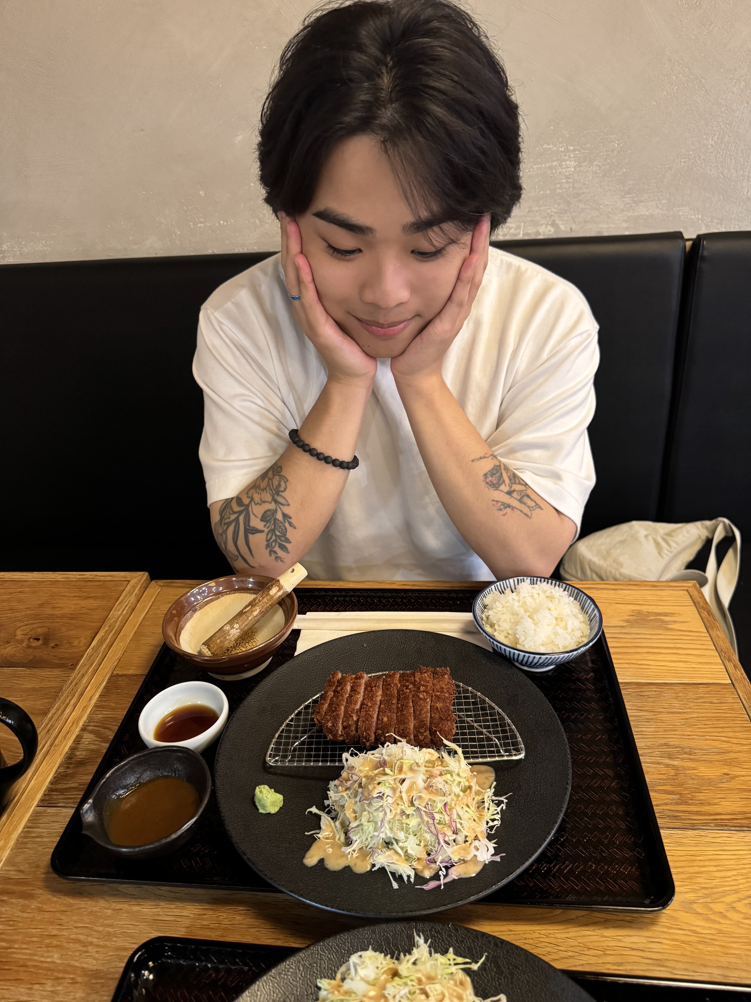 A young man with dark hair and tattoos on his arms, sitting at a table in a restaurant, looking down with his hands on his face, surrounded by various dishes including rice, salad, and a grilled meat dish.