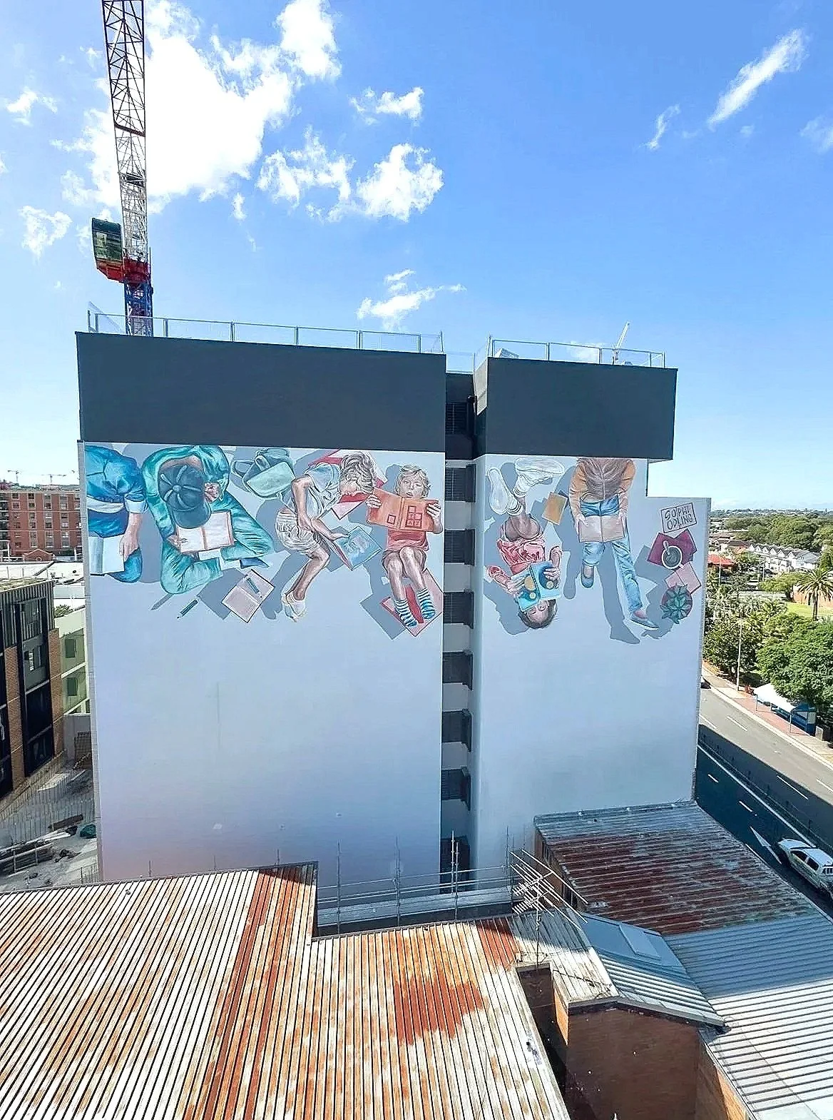 A tall building with a large mural featuring animated characters on its side, under a blue sky with scattered clouds. There are rooftops and streets visible in the foreground.