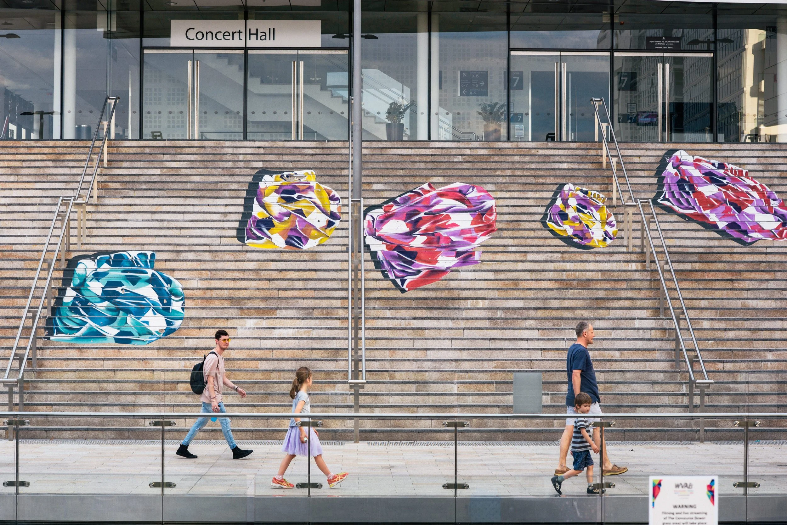 Modern performing arts center with large white geometric facade, glass windows, and steps with colorful umbrellas, people walking, and a clear sky.