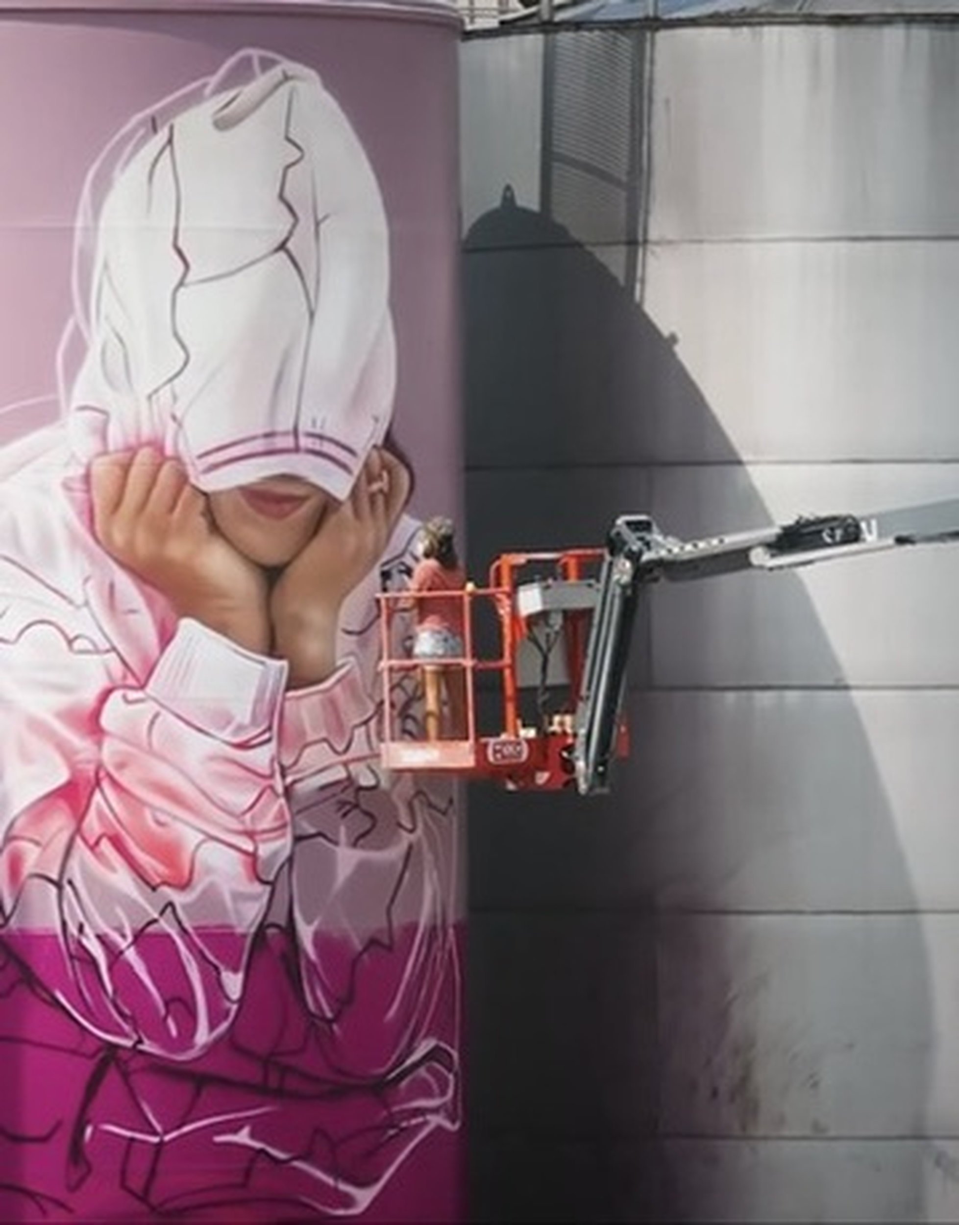 A person wearing a pink hoodie with a white graphic design, resting their chin on their hands. They are holding their face with both hands and are next to a vending machine or some kind of metallic structure. The person is partially obscured by a white paper cutout or illustration of a face mask or head covering with black outlines.