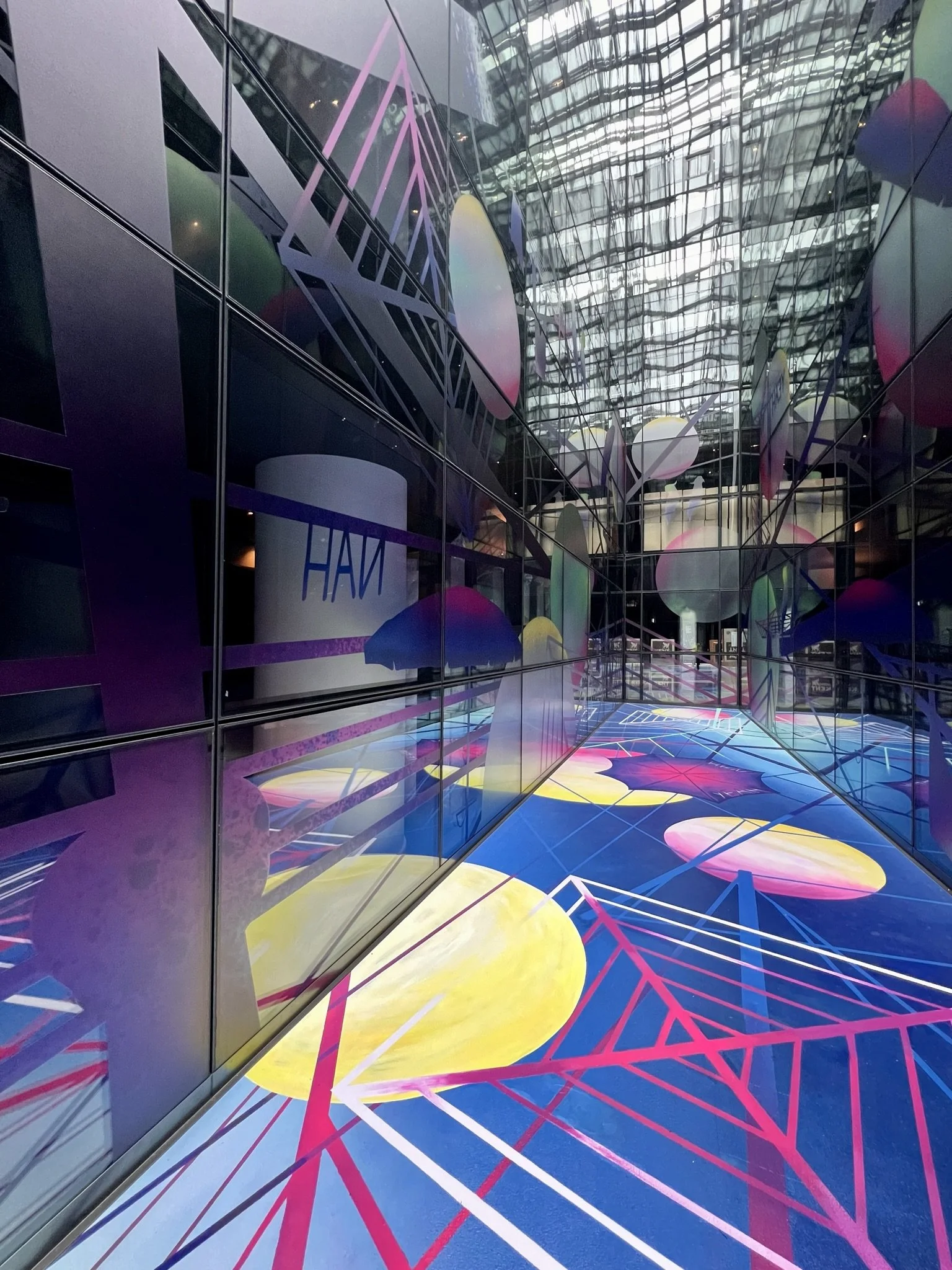 Colors and lines create an abstract, digitally inspired mosaic of shapes on a glass building interior wall, in an urban setting with reflections and patterns.