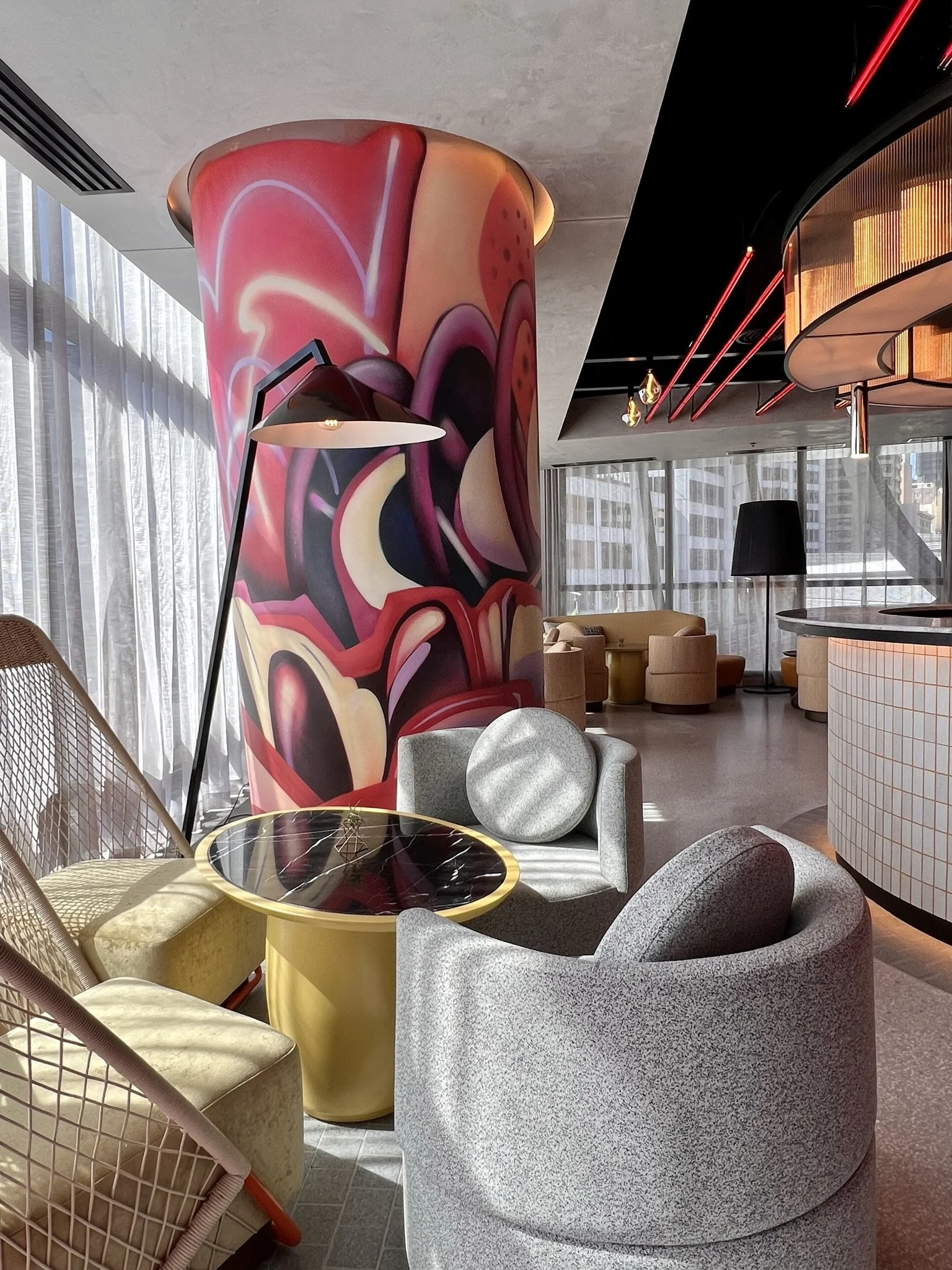 Interior of a modern restaurant with vibrant decor, including a large painted column with abstract floral artwork, red and yellow neon light accents on the ceiling, patterned black and white flooring, and seating areas with tables and chairs.