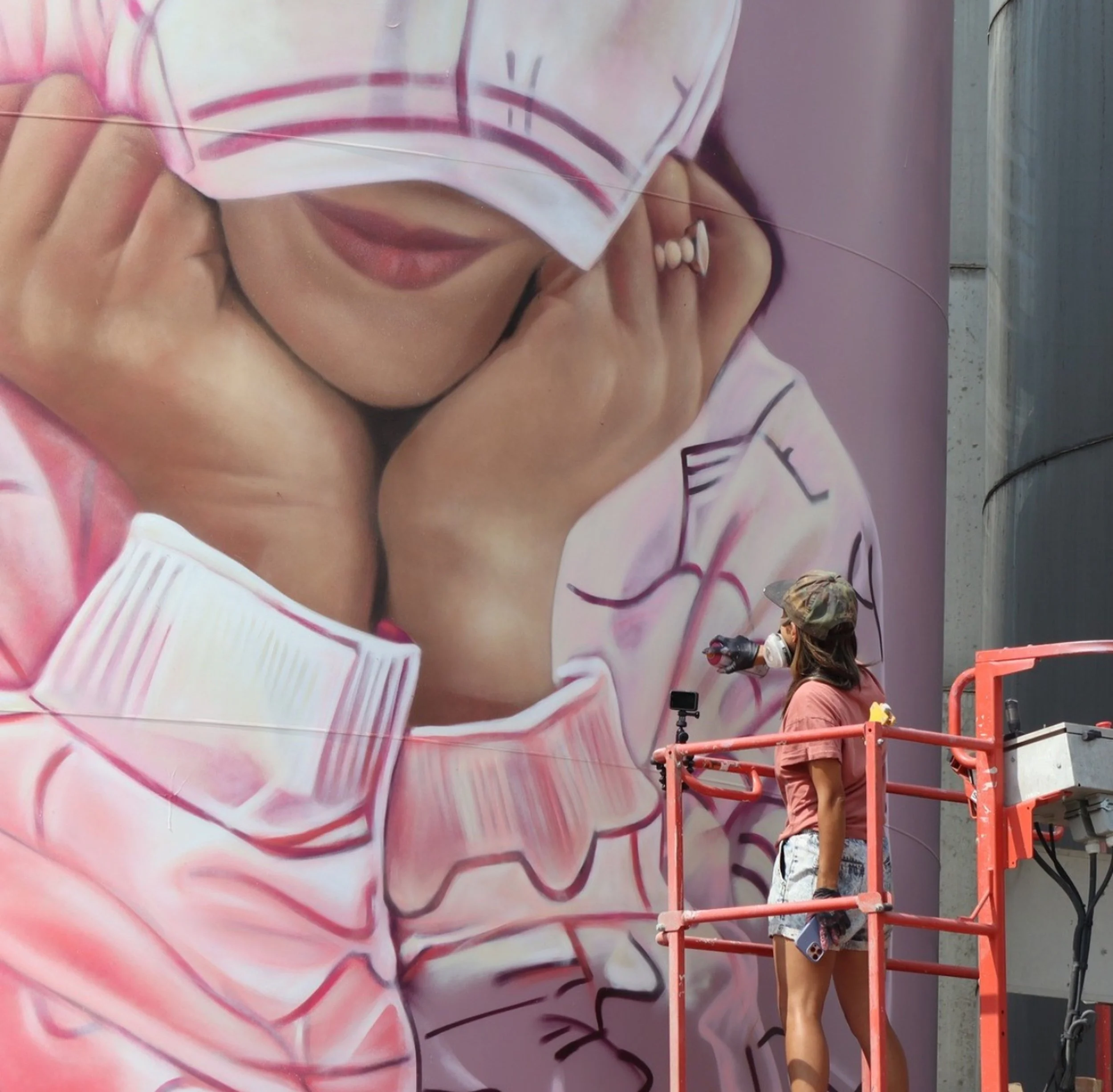 Workers on a scaffolding painting a large mural of a woman's face on a wall. The mural features the woman's lips and part of her eye visible, with a pink background.