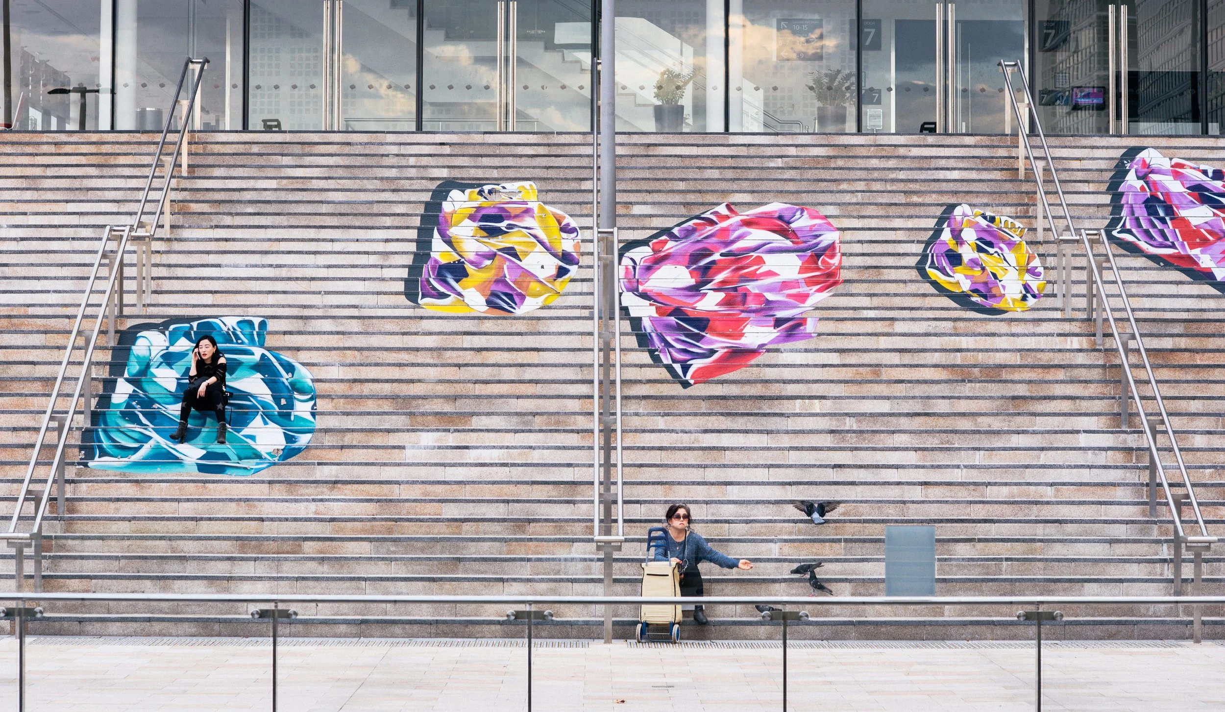 People sitting and standing on a wide outdoor staircase decorated with colorful floral murals, in front of a modern glass building.